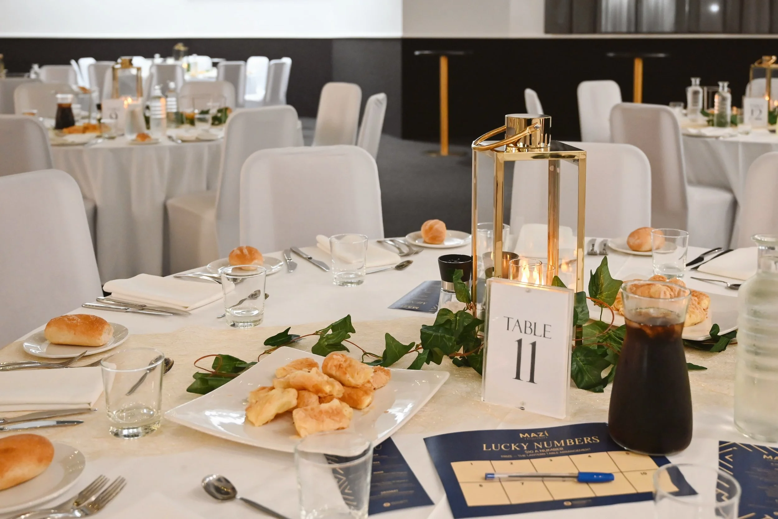 Elegant banquet dining table with white tablecloth, gold geometric lantern centerpiece, white chairs, glassware, bread rolls, and a drink, set for an event.
