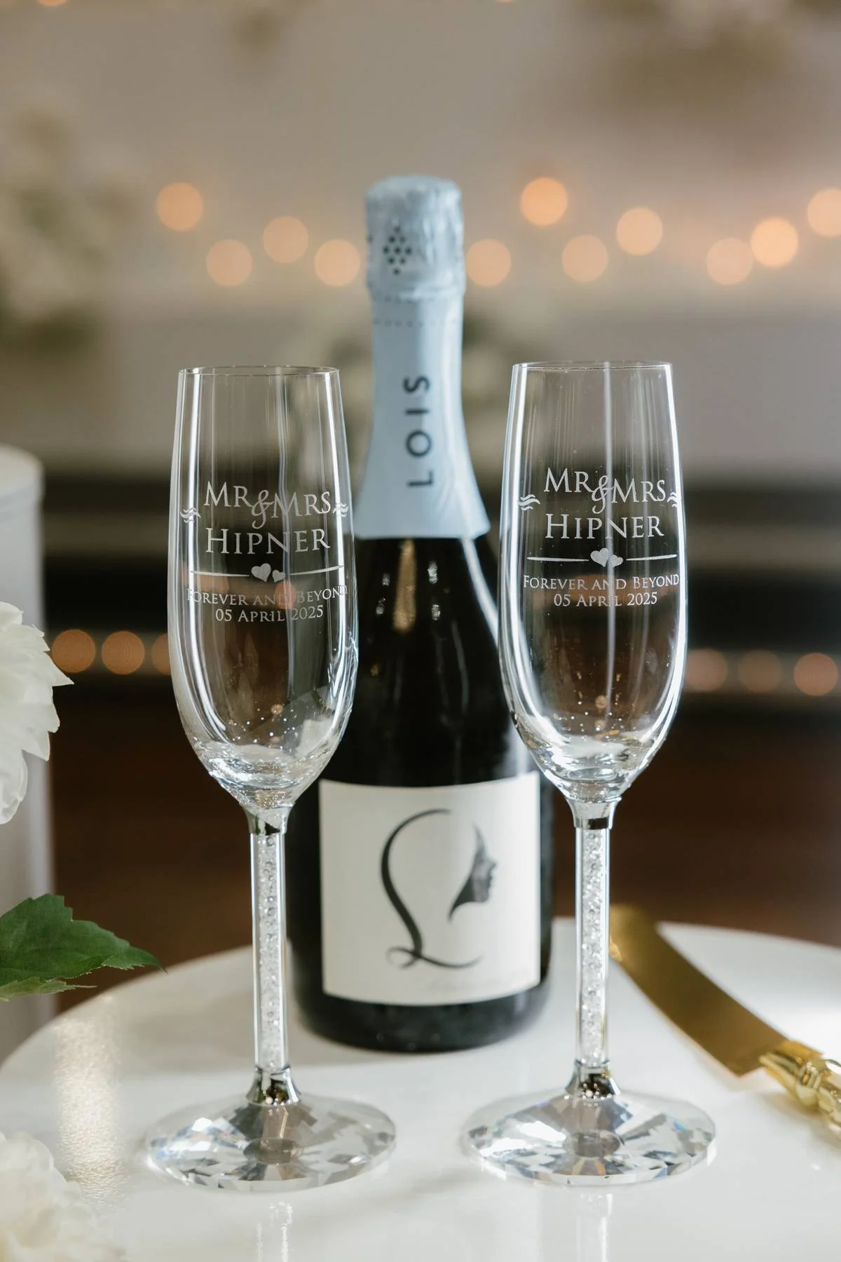 Two champagne flutes with engraved wedding message, a bottle of sparkling wine in the background with a blurred bokeh light effect, and a white flower partially visible on the left at Donato the venue in Adelaide