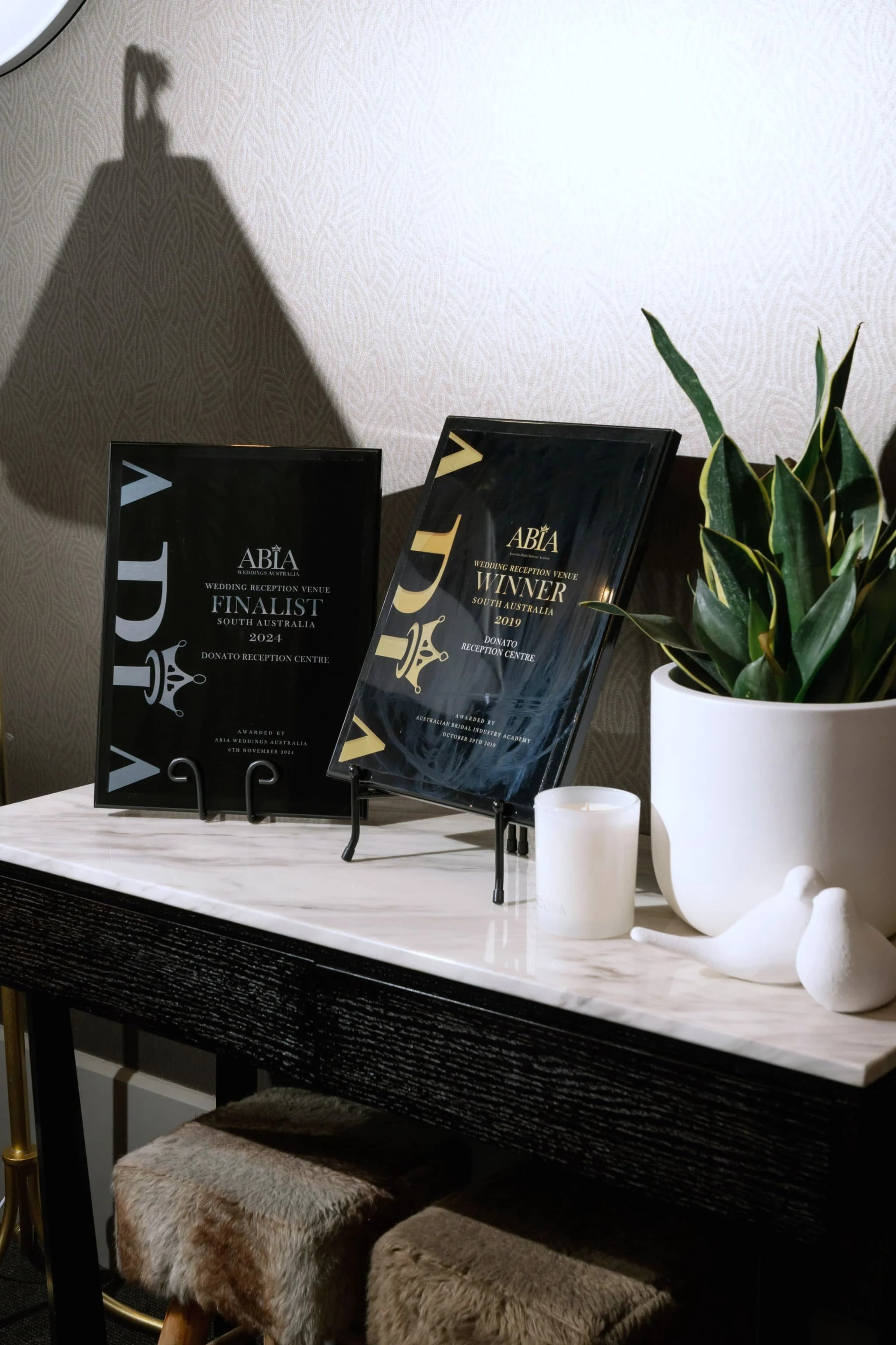 Two awards plaques, a plant in a white pot, candles, and faux fur-covered stools on a white marble table, with a beige patterned wall in the background.