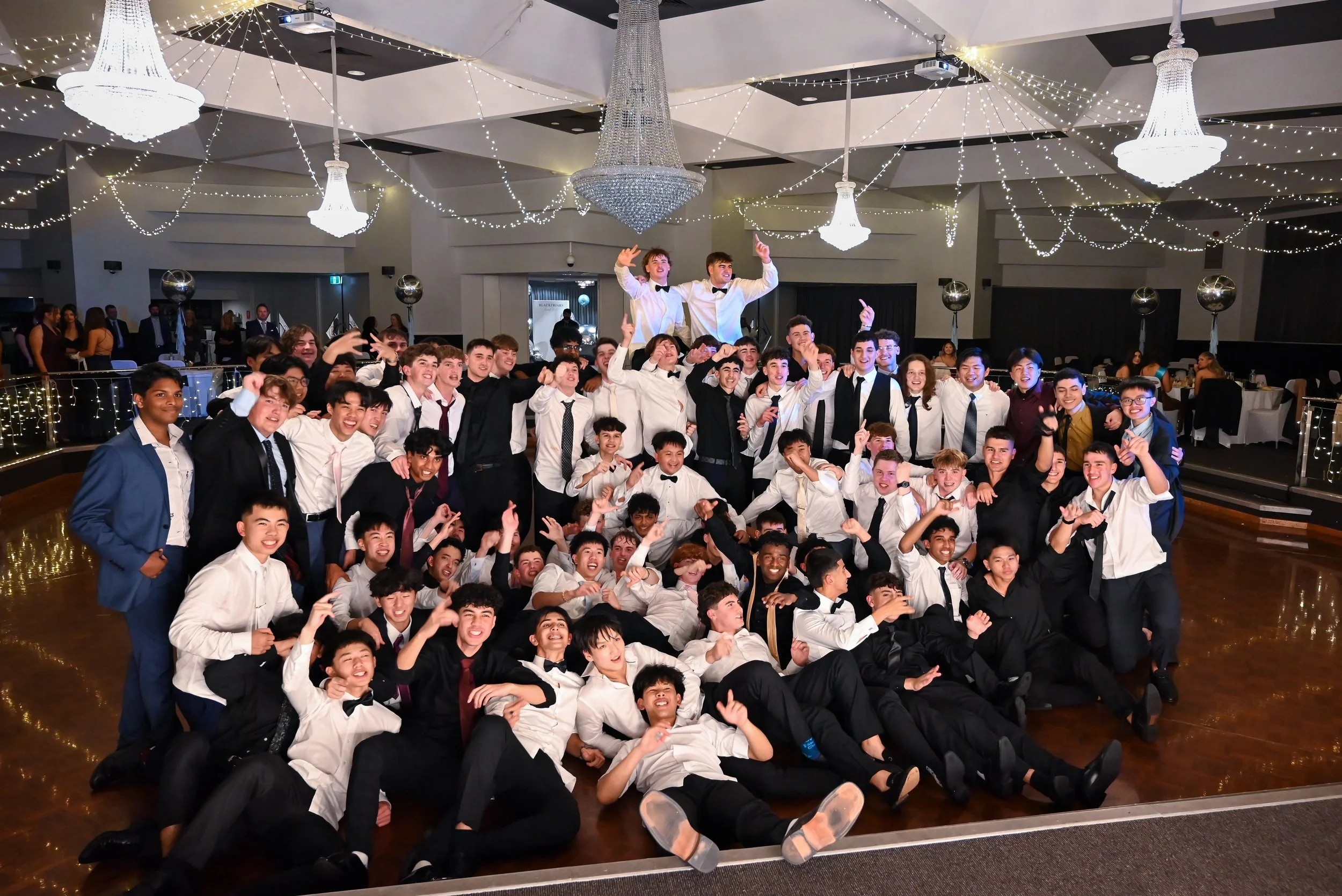 A large group of young people dressed in formal attire, celebrating at a banquet hall decorated with chandeliers and string lights, with two boys being lifted on others' shoulders.