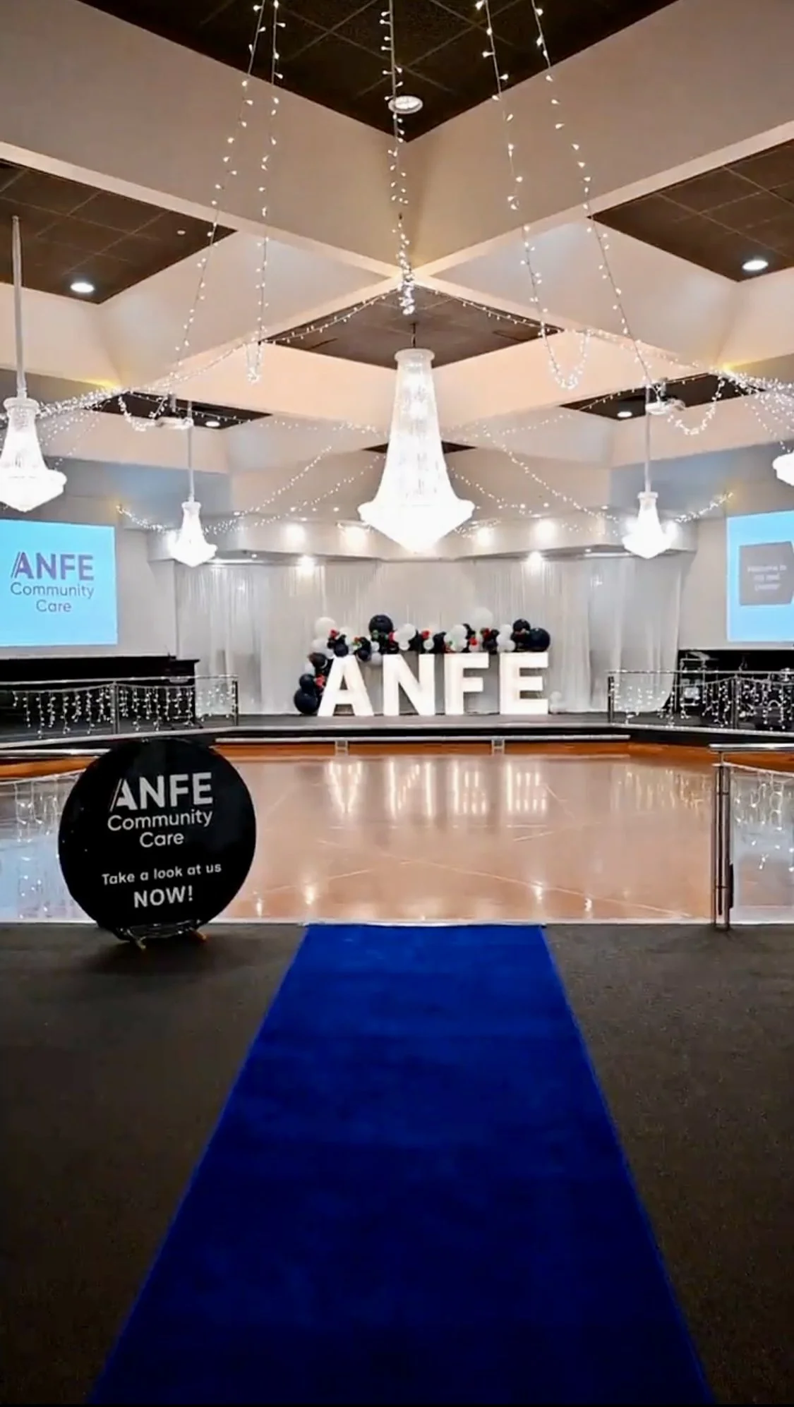 Event hall decorated with white curtains, chandeliers, string lights, black and white floral arrangements, and large illuminated letters spelling 'ANFE' at the center. There is a blue carpet leading to the stage with a sign on a black balloon that sa
