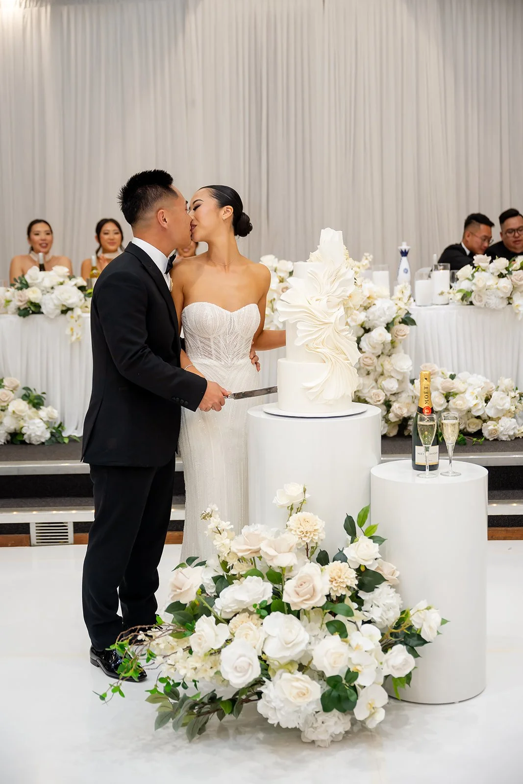A bride and groom kiss while cutting a wedding cake at their wedding reception, with wedding party and guests seated at a decorated table in the background.
