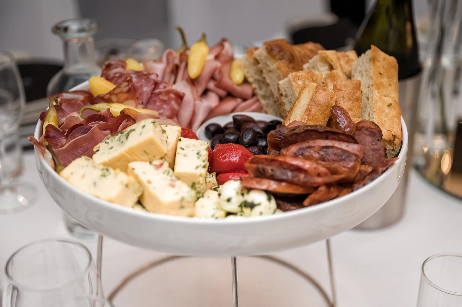 Charcuterie board with cheeses, meats, bread, olives, and grapes, served on a white platter at a social gathering.