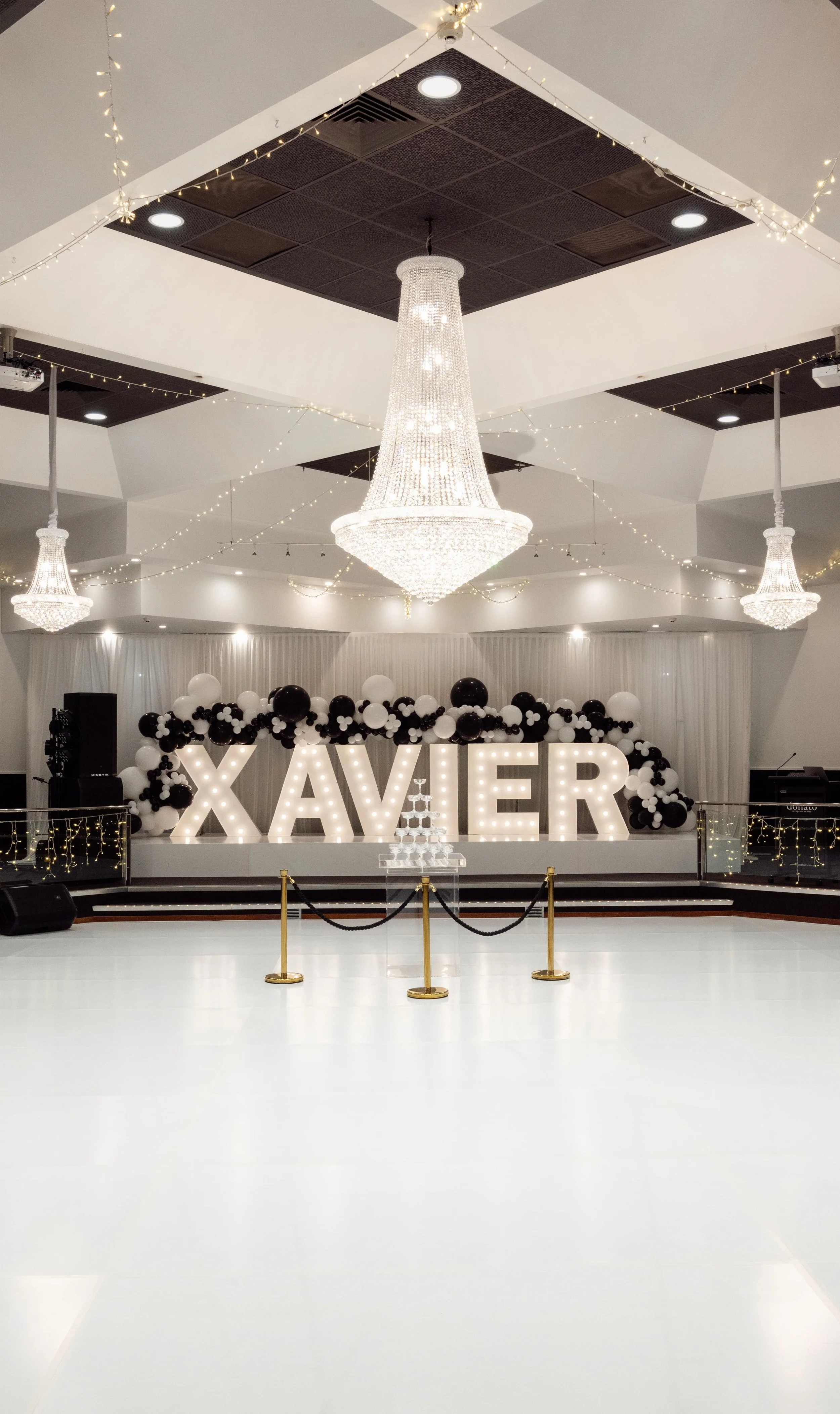Event hall decorated with large illuminated letters spelling 'XAVIER,' black and white balloons, and a tiered champagne tower on a stage. Multiple chandeliers and string lights adorn the ceiling.