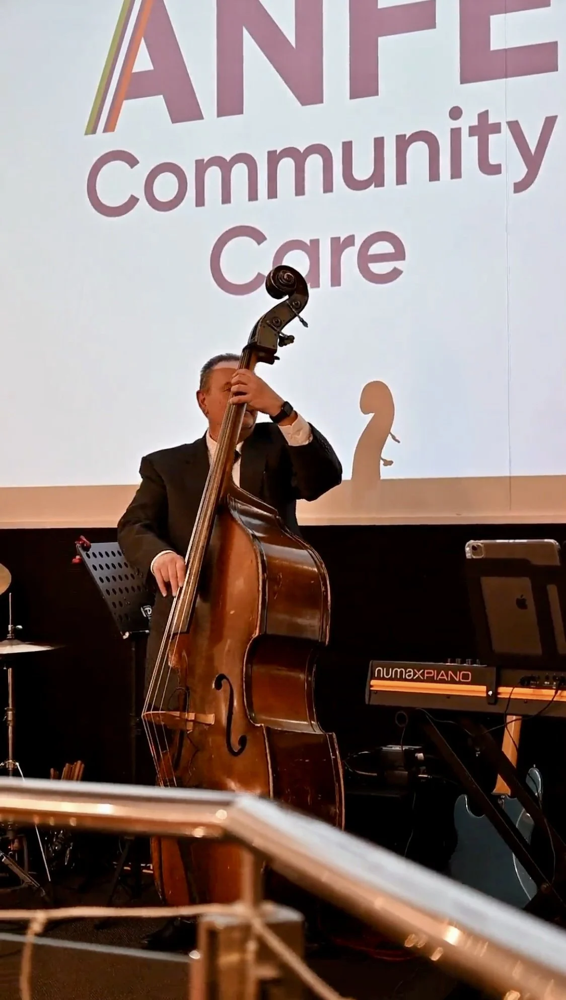 A man in a black suit playing a double bass on stage during a community event, with a large screen in the background displaying the words 'Community Care' and part of the word 'FAMILY'.