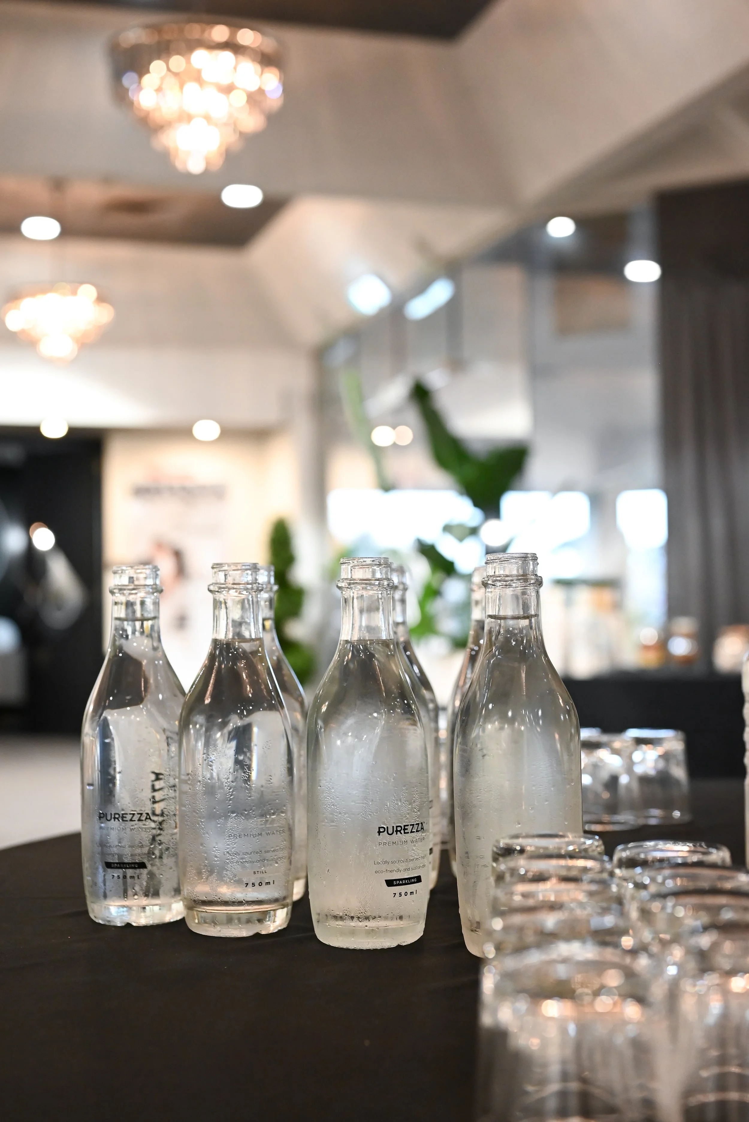Row of clear bottled water labeled 'PUREZZA' on a dark table in a well-lit modern restaurant or event space with chandeliers and large windows.