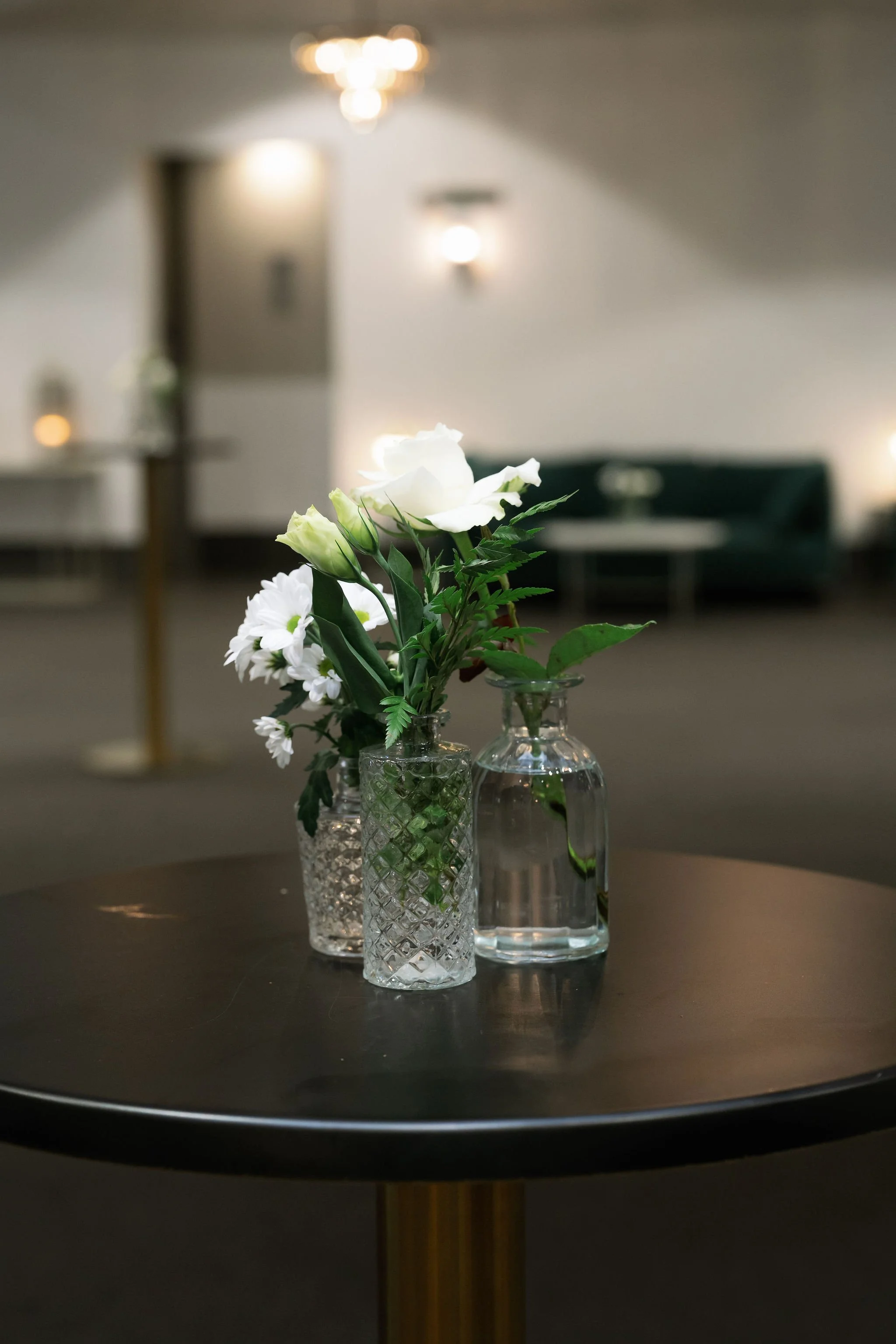 Three glass vases with white flowers on a table, blurred background of a living room with lights and a green couch.