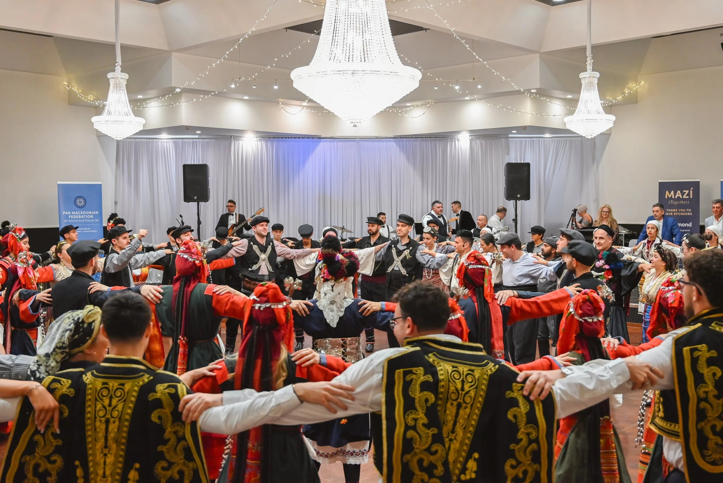 A large group of people dressed in traditional clothing are dancing in a circle at an indoor cultural event. The background features a stage with musical instruments and musicians, and the room is decorated with elegant chandeliers and string lights.