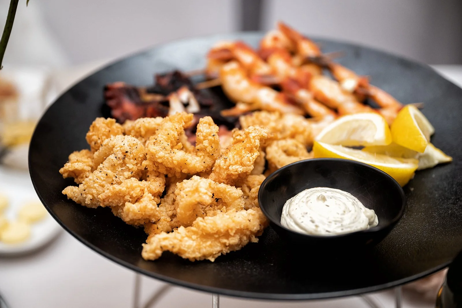 Fried calamari, shrimp skewers, lemon wedges, and dipping sauce on a black serving plate.