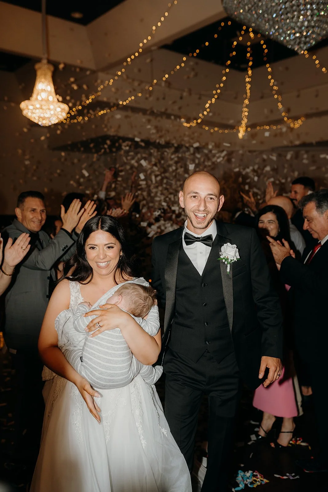 The Bride and Groom with their infant and people are clapping with a smile.