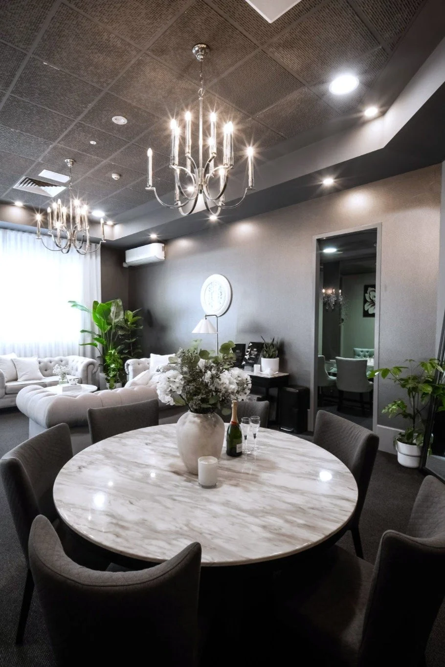 Elegant dining and lounge area with a round marble table, floral centerpiece, champagne bottle, and comfortable seating, illuminated by chandeliers and ceiling lights.