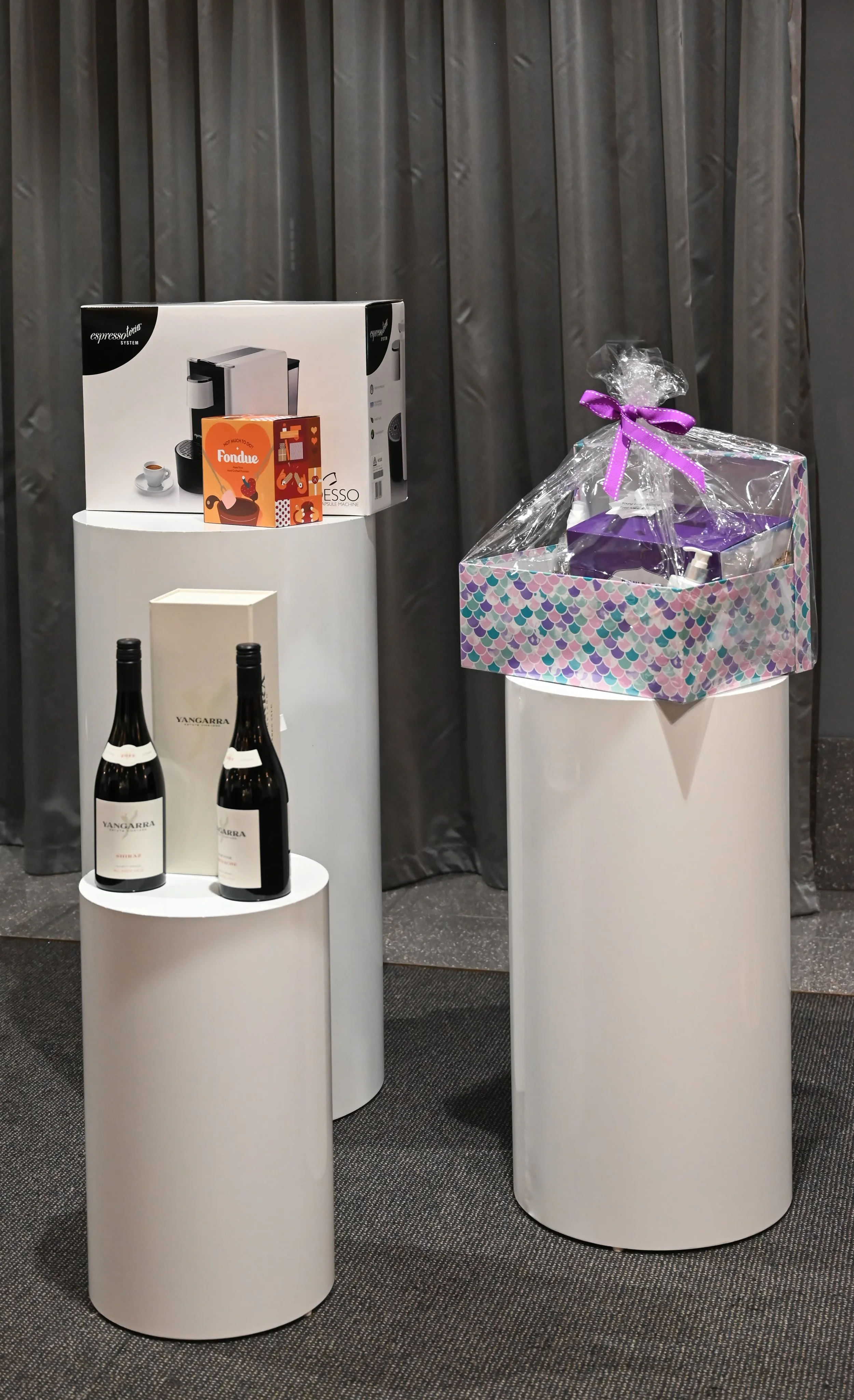 Display of wine bottles on white cylindrical pedestals, a gift basket with a purple ribbon, and a coffee machine box against a gray curtain backdrop.