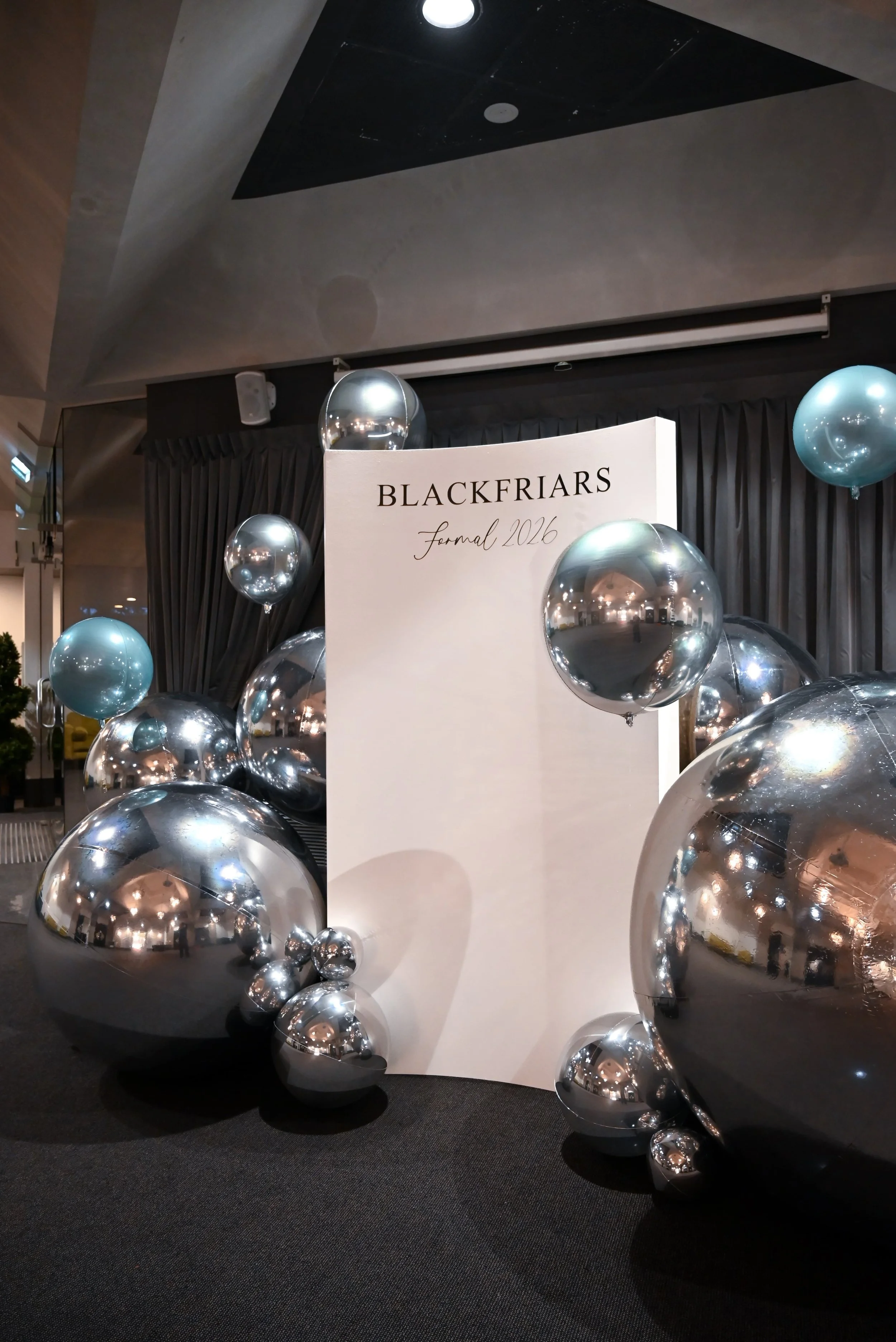 A display featuring numerous silver balloons surrounding a white sign with the text 'Blackfriars Formal 2026'. The balloons are reflective, and the display is set against a dark curtain backdrop in an indoor setting.