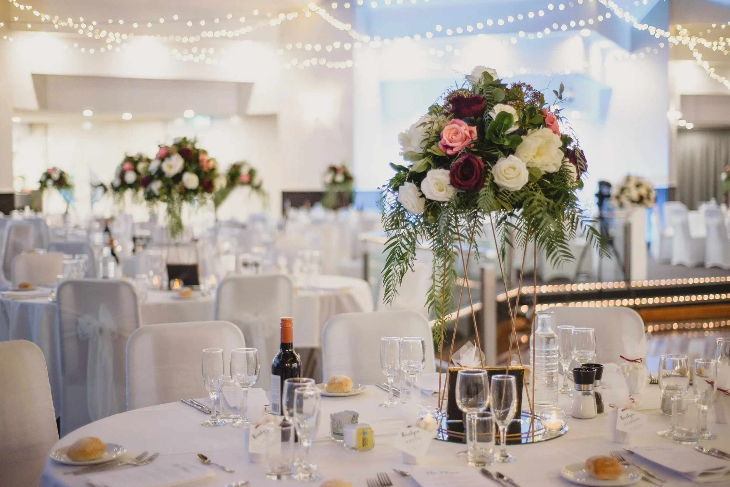Elegant banquet hall decorated for a wedding reception with white tablecloths, floral centerpieces, and string lights overhead.