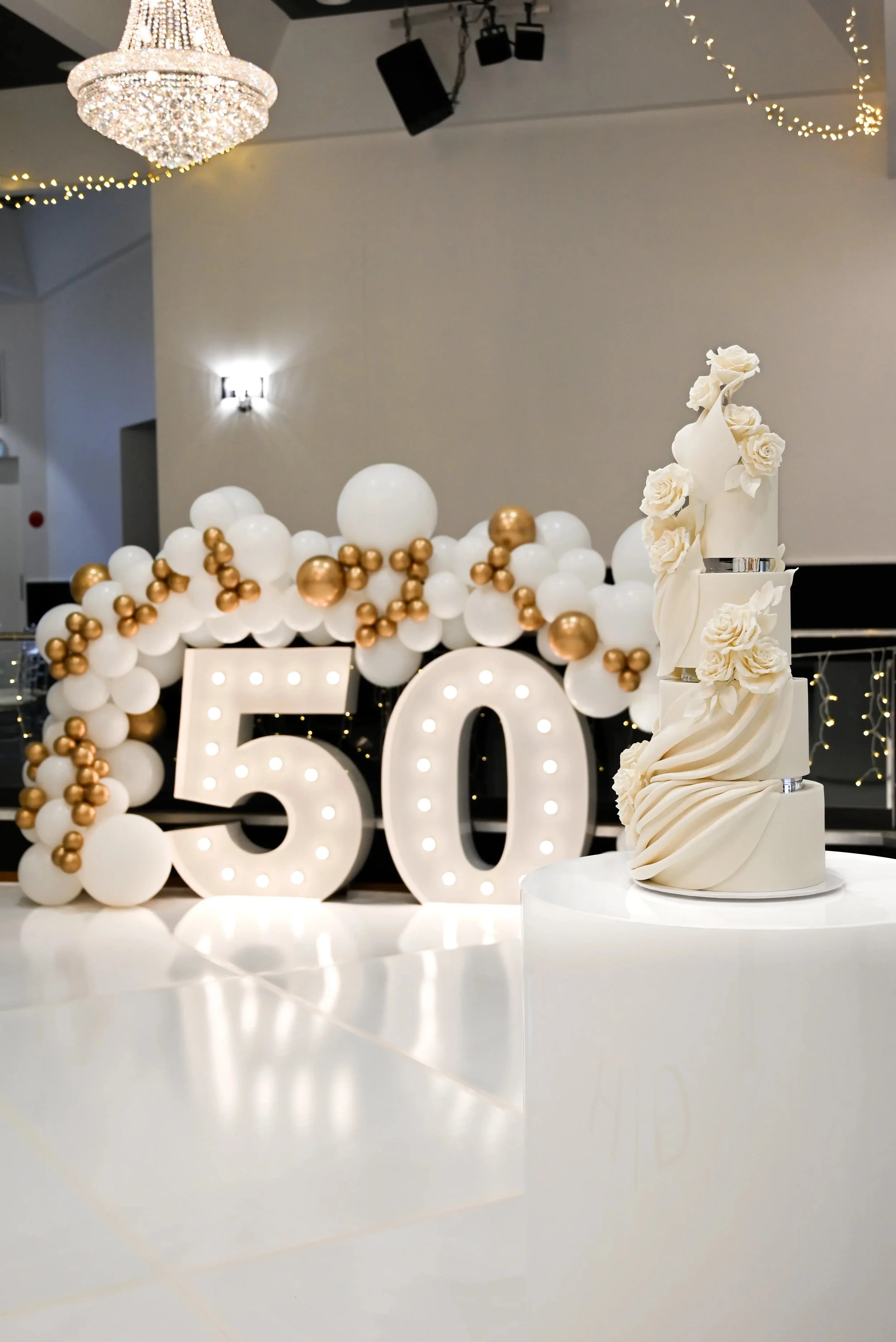 A decorative celebration display featuring a large illuminated number five, white balloons with gold accents, and a white sculpted cake adorned with white roses and flowing fabric-like details.