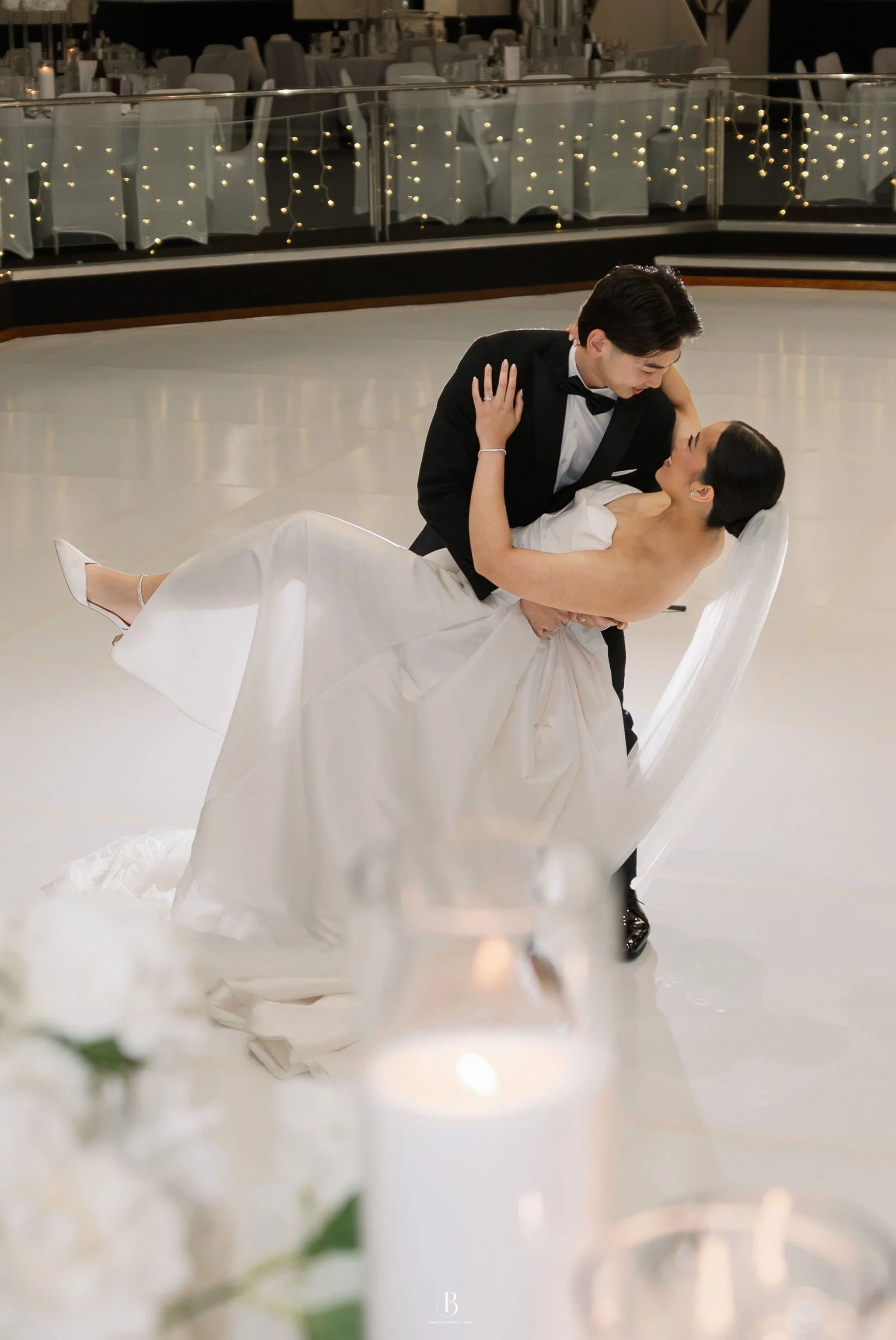 A bride and groom dancing at their wedding reception. The groom is wearing a black tuxedo and the bride is in a white wedding gown. They are looking into each other's eyes, with the groom holding the bride while she leans back. The background feature