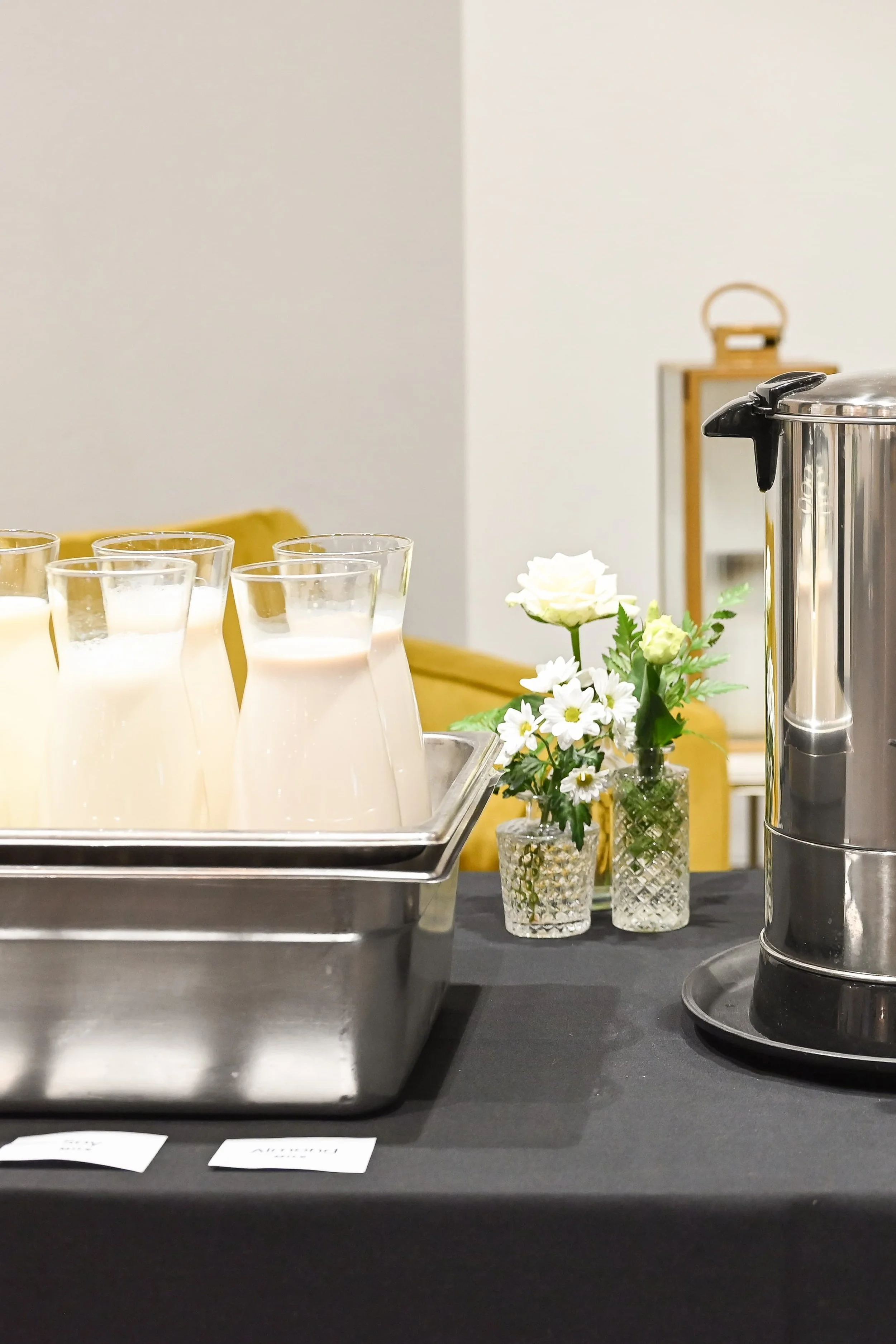 A buffet table with glasses of milk, small white flowers in glass vases, a large silver coffee urn, and a yellow chair in the background.