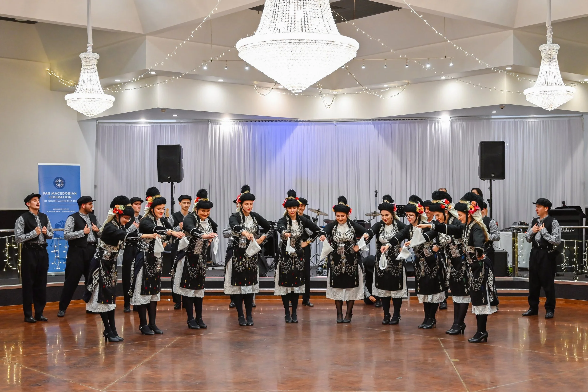 A group of people dressed in traditional clothing performing a dance on stage at an indoor event. There are chandeliers and white curtains in the background, and a banner for the Pan Macedonian Federation is visible on the left side.