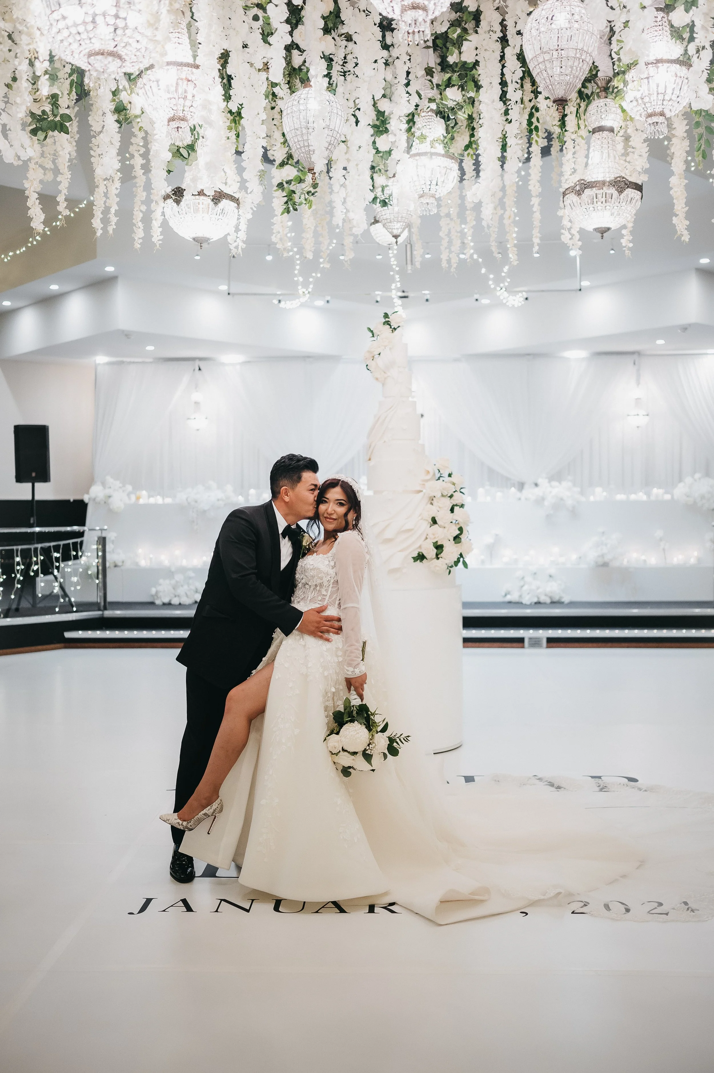 A bride and groom pose intimately in the center of a wedding venue decorated with white flowers and hanging lanterns. The bride is wearing a long lace wedding gown, and the groom is in a black tuxedo. The floor has the month and year 'January 2023' w