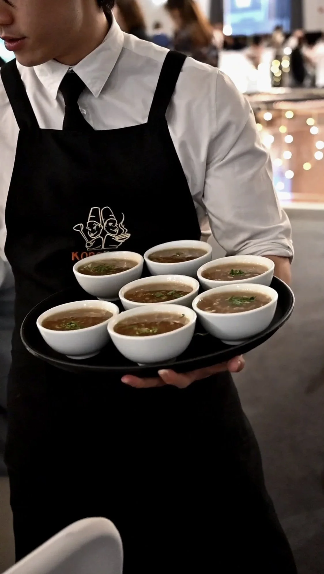 A waitress in a white shirt, black tie, and black apron holding a tray of eight bowls of soup in a restaurant setting.