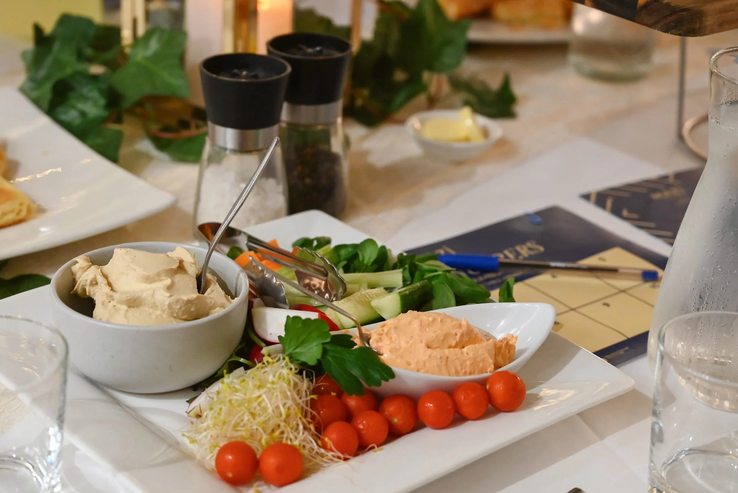 A table set with various appetizers, including cherry tomatoes, sliced vegetables, and dips, with condiments and salt and pepper shakers in the background.