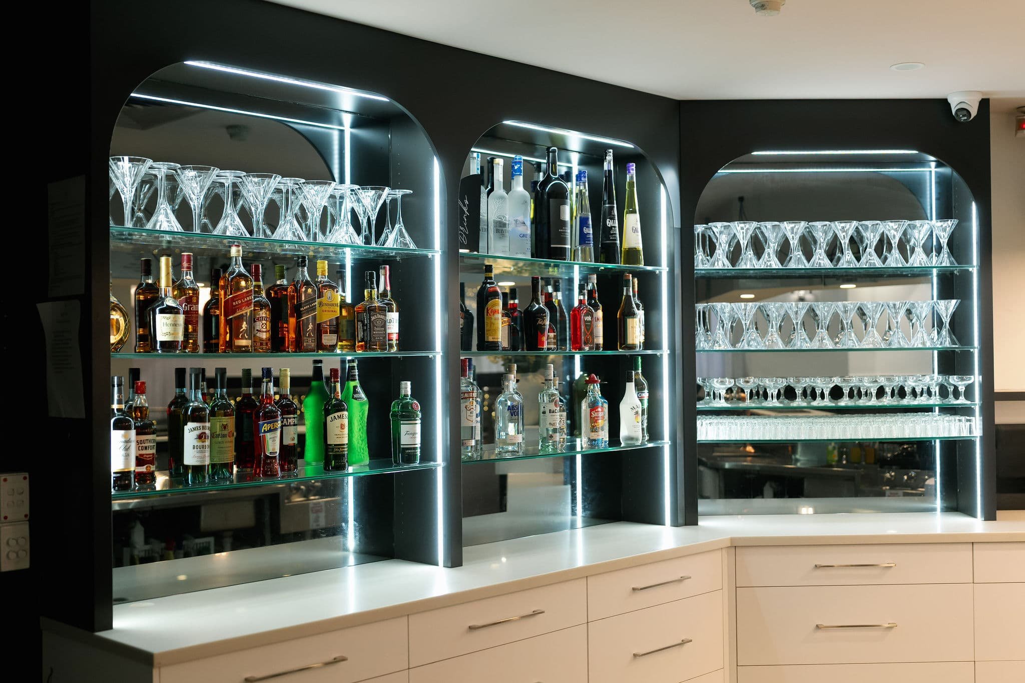 A modern bar shelf with illuminated glassware and bottles of liquor on glass shelves at Donato the venue in Adelaide