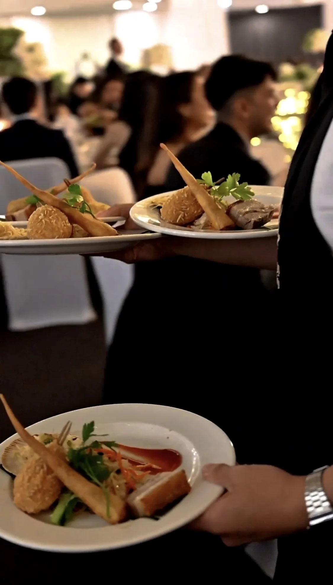 People in formal attire enjoying a banquet with waitstaff serving plates of food at a celebration or event.