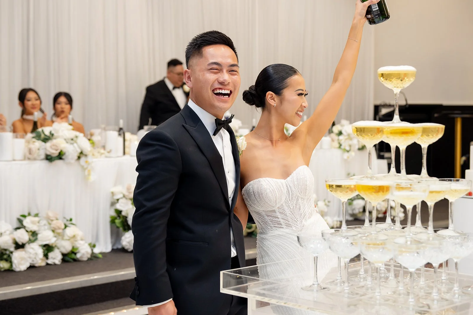Happy bride and groom at their wedding reception, standing near a champagne tower, with guests and floral decorations in the background at Donato reception centre in Adelaide