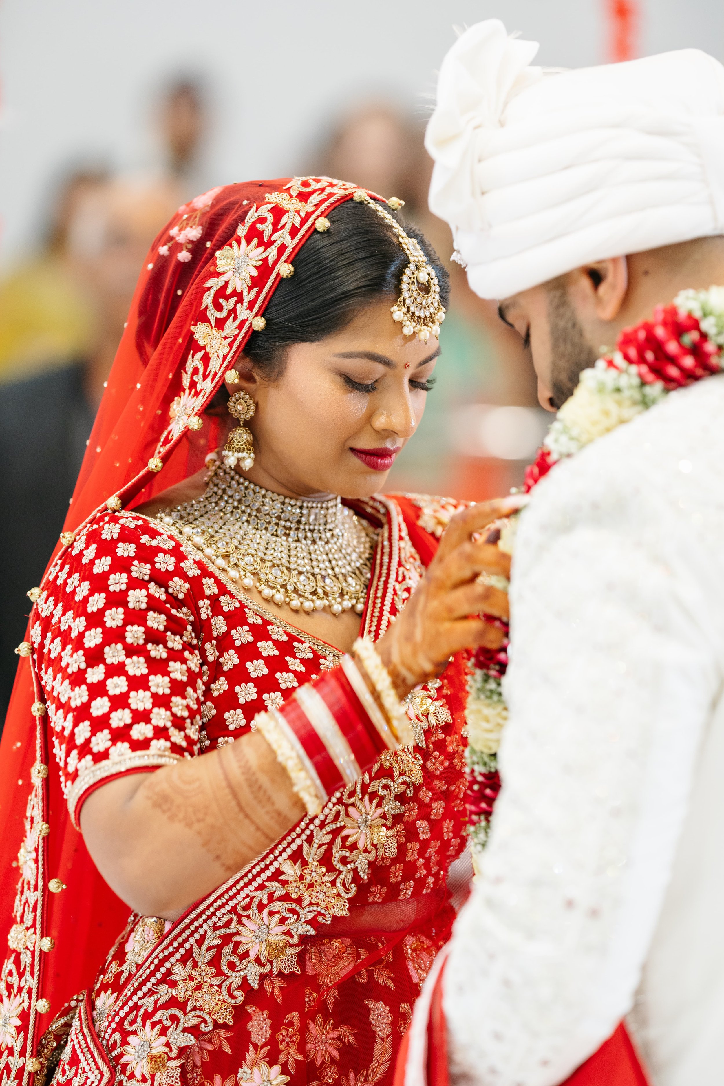 A bride and groom in traditional Indian wedding attire during a wedding ceremony. The bride is dressed in a red saree with gold and pearl jewelry, and the groom is wearing a white sherwani with a turban.