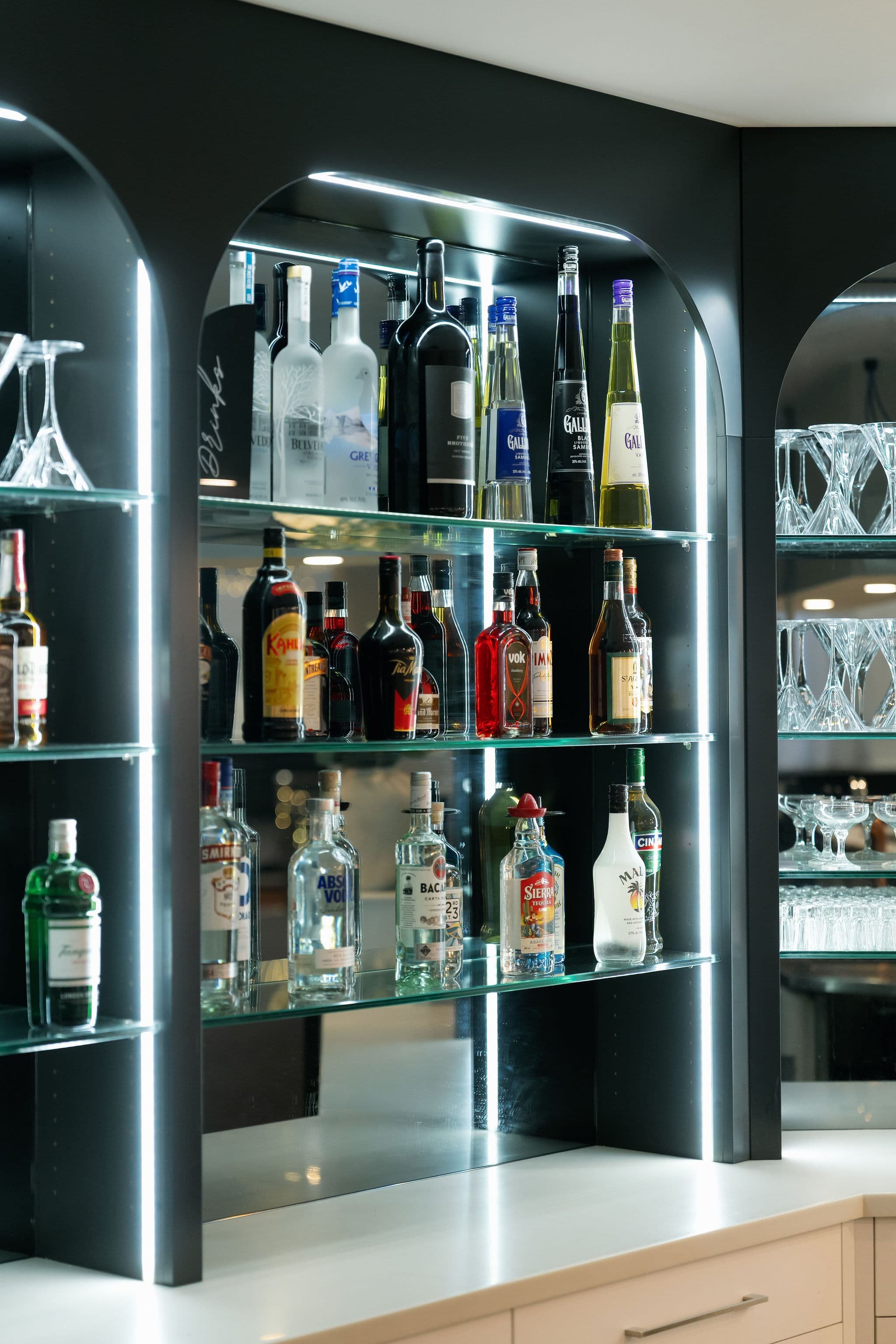 A modern bar with glass shelves holding various liquor bottles and glassware, illuminated by sleek LED lighting.