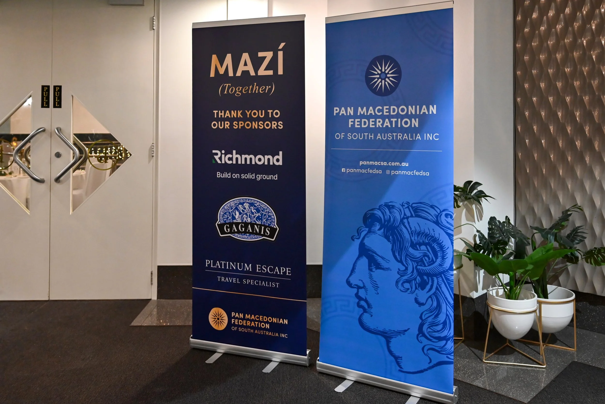 Two vertical banners displayed indoors. The left banner is dark blue with the word 'MAZI (Together)' in large letters, and mentions 'Thank you to our sponsors' with logos for Richmond and Gaganis. The right banner is light blue with a stylized lion h