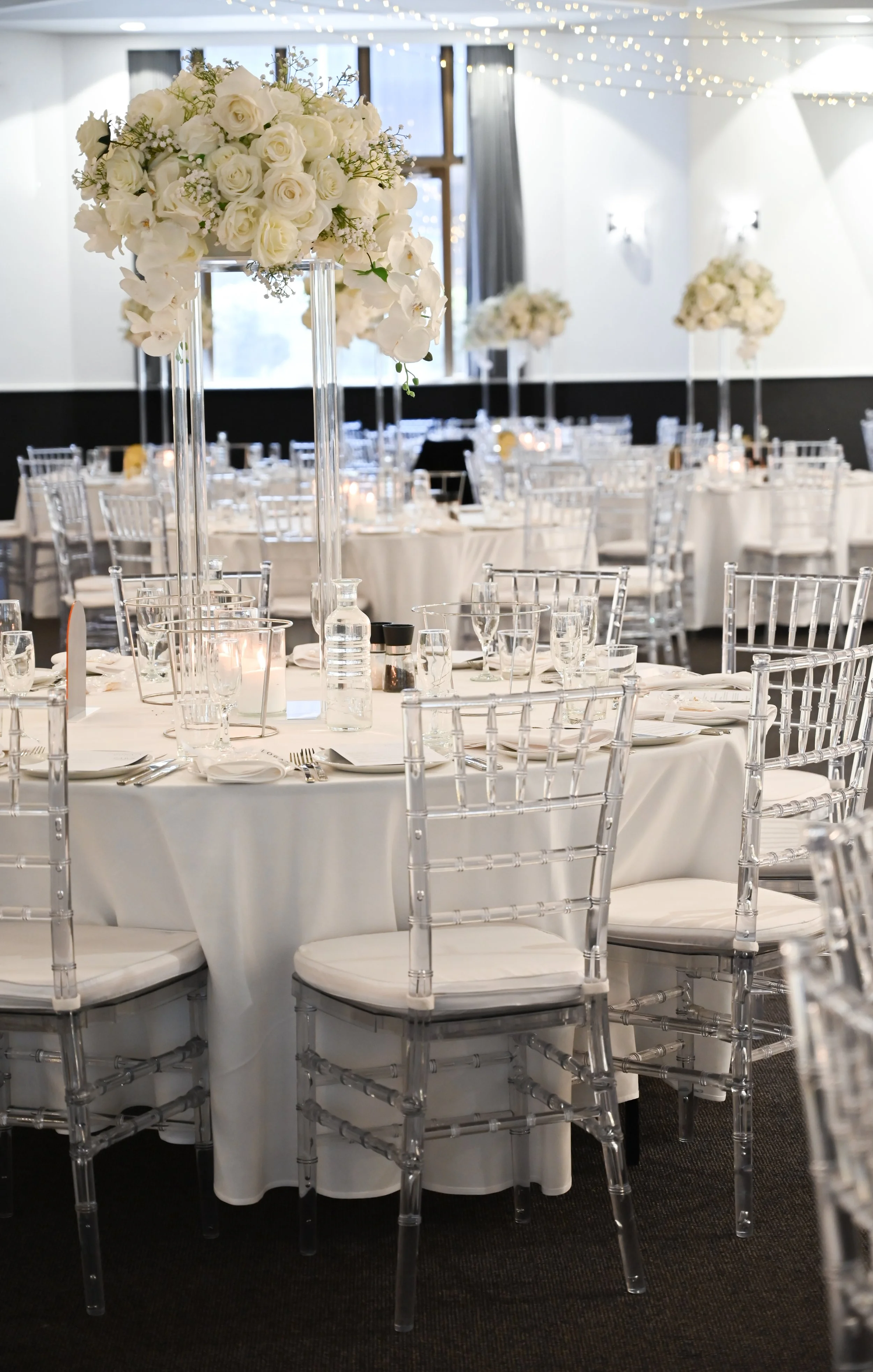 Elegant event space decorated with white floral arrangements on tall glass stands, round tables with white tablecloths, and clear acrylic chairs. Soft lighting with string lights and wall sconces add ambiance.