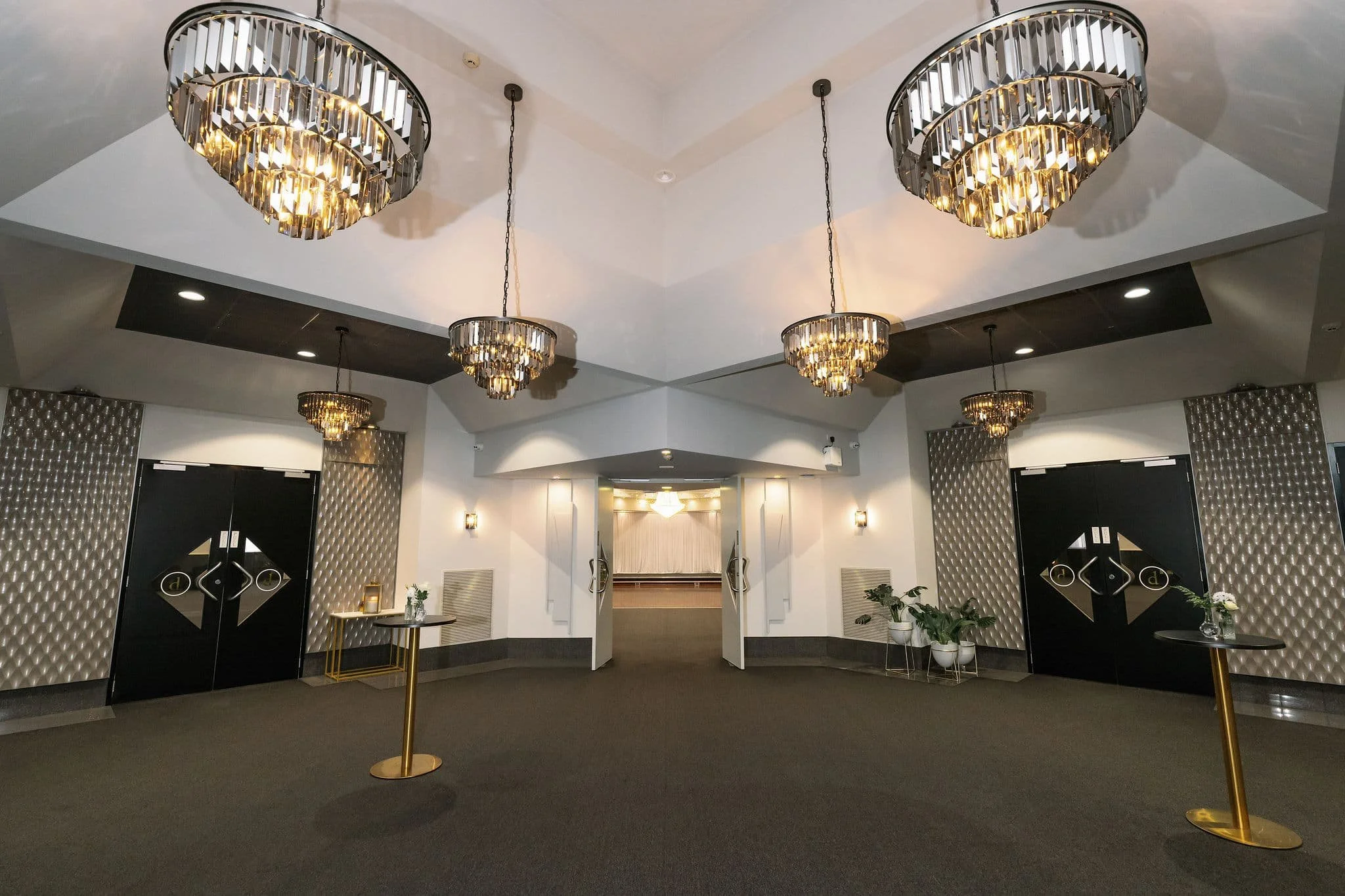Lobby with multiple large, modern chandeliers hanging from the ceiling, dark carpeted floor, decorative textured wall panels, small tables with plants, and double doors with geometric designs at Donato the venue in Adelaide