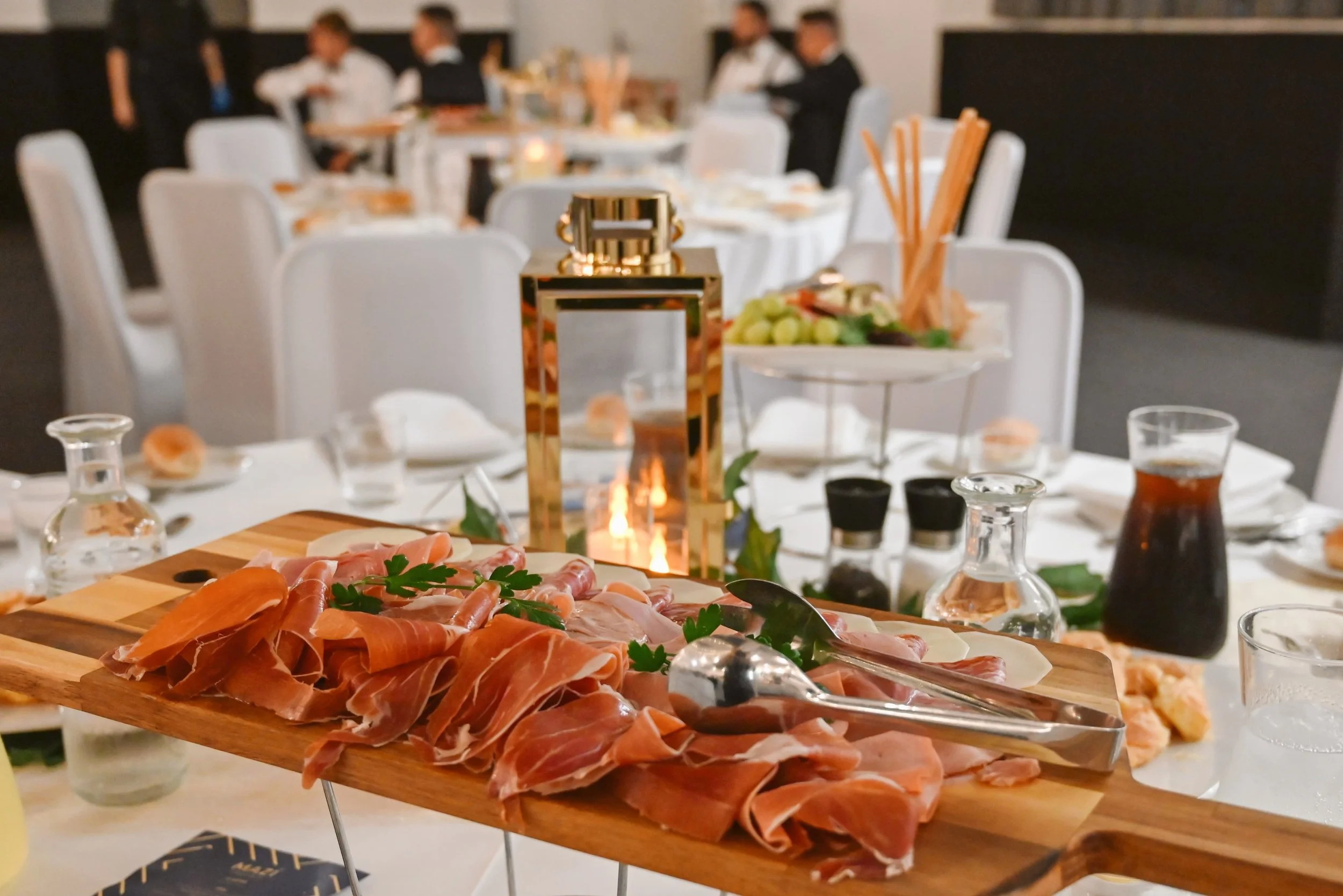Close-up of a wooden platter with various sliced cured meats garnished with parsley, placed on a table at a restaurant. In the background, there are blurred tables with white tablecloths, glassware, and a lit candle, with people standing and talking.