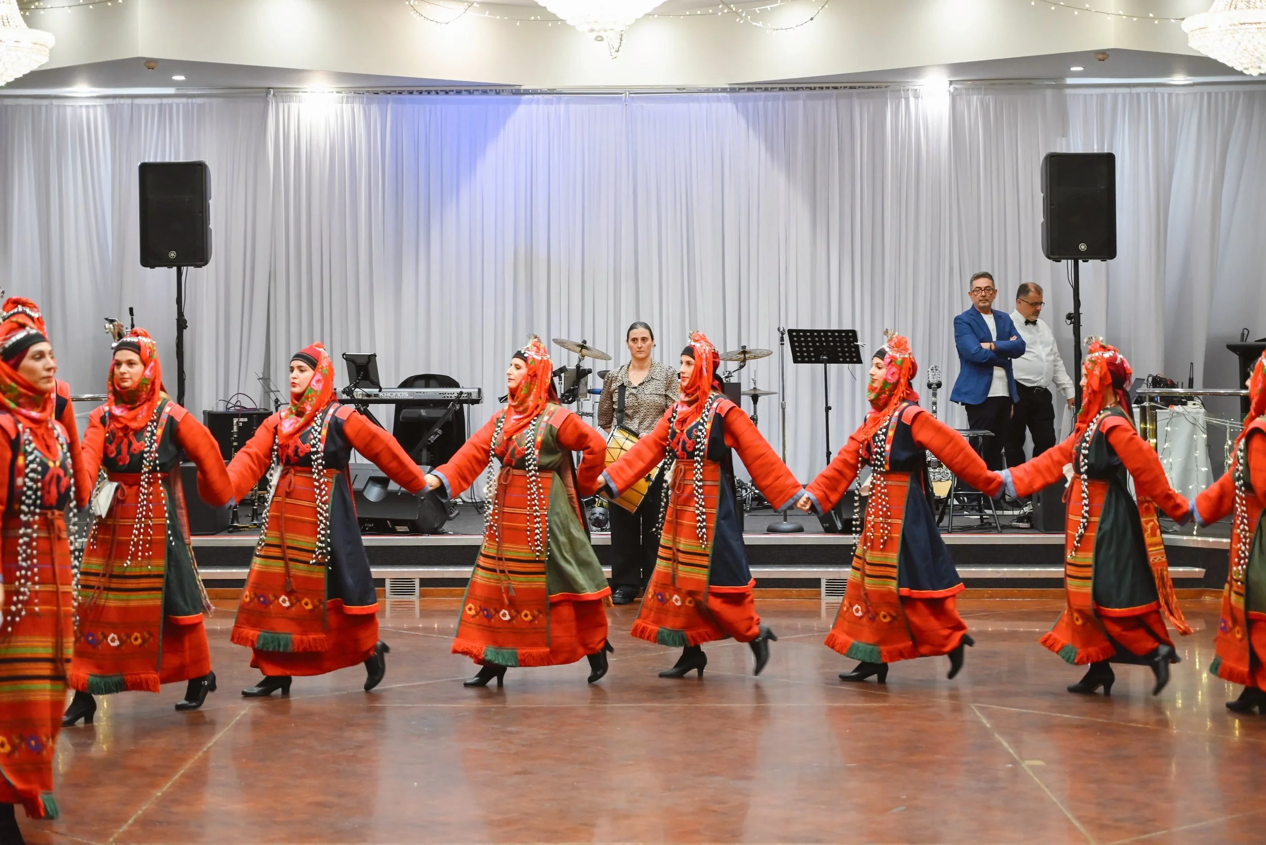 A group of women dressed in traditional, colorful folk costumes are performing a dance indoors. They are holding hands and moving in a circle. In the background, there are musicians, including a woman with a drum and men standing near sound equipment