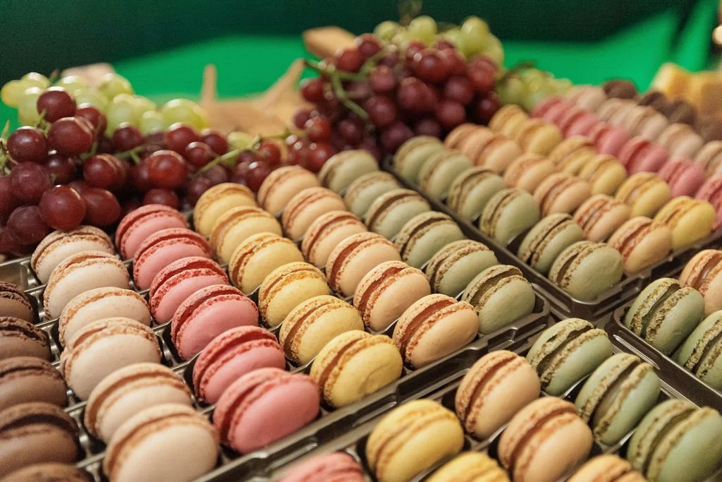 Assorted pastel-colored macarons arranged on display with green and red grapes in the background.