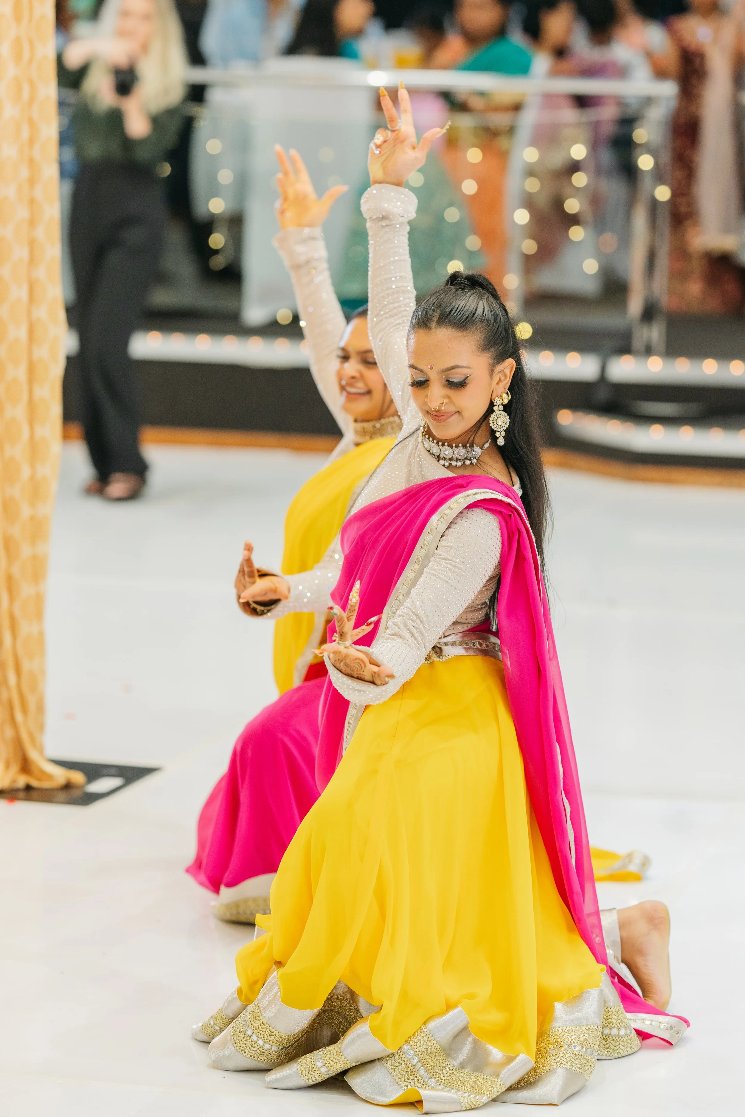 Two women in traditional Indian attire performing a dance, kneeling with hands raised, in front of a blurred audience.