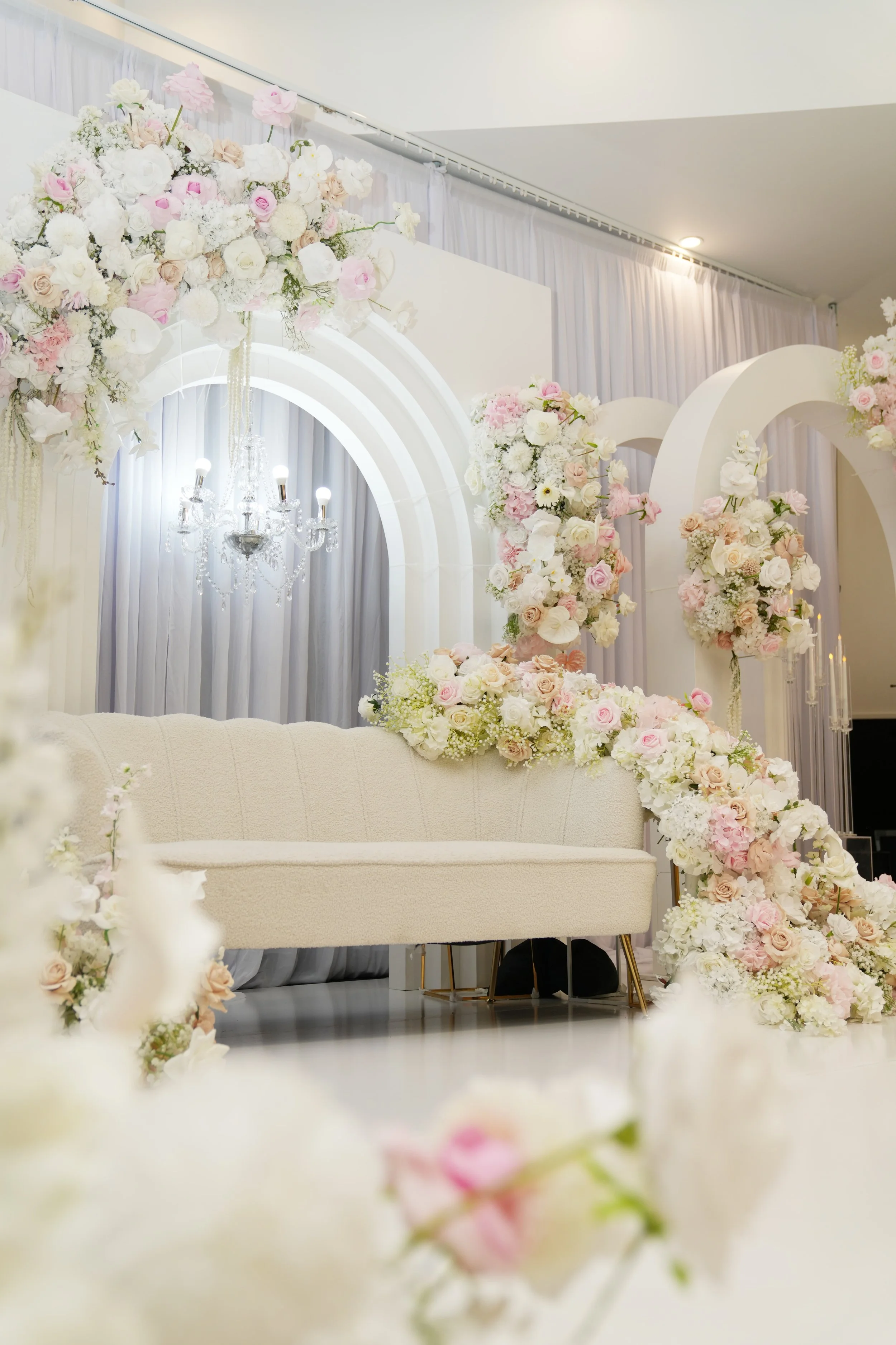 Elegant wedding stage decorated with white and pink flowers, a beige sofa, a crystal chandelier, and white drapes in the background at Donato the venue