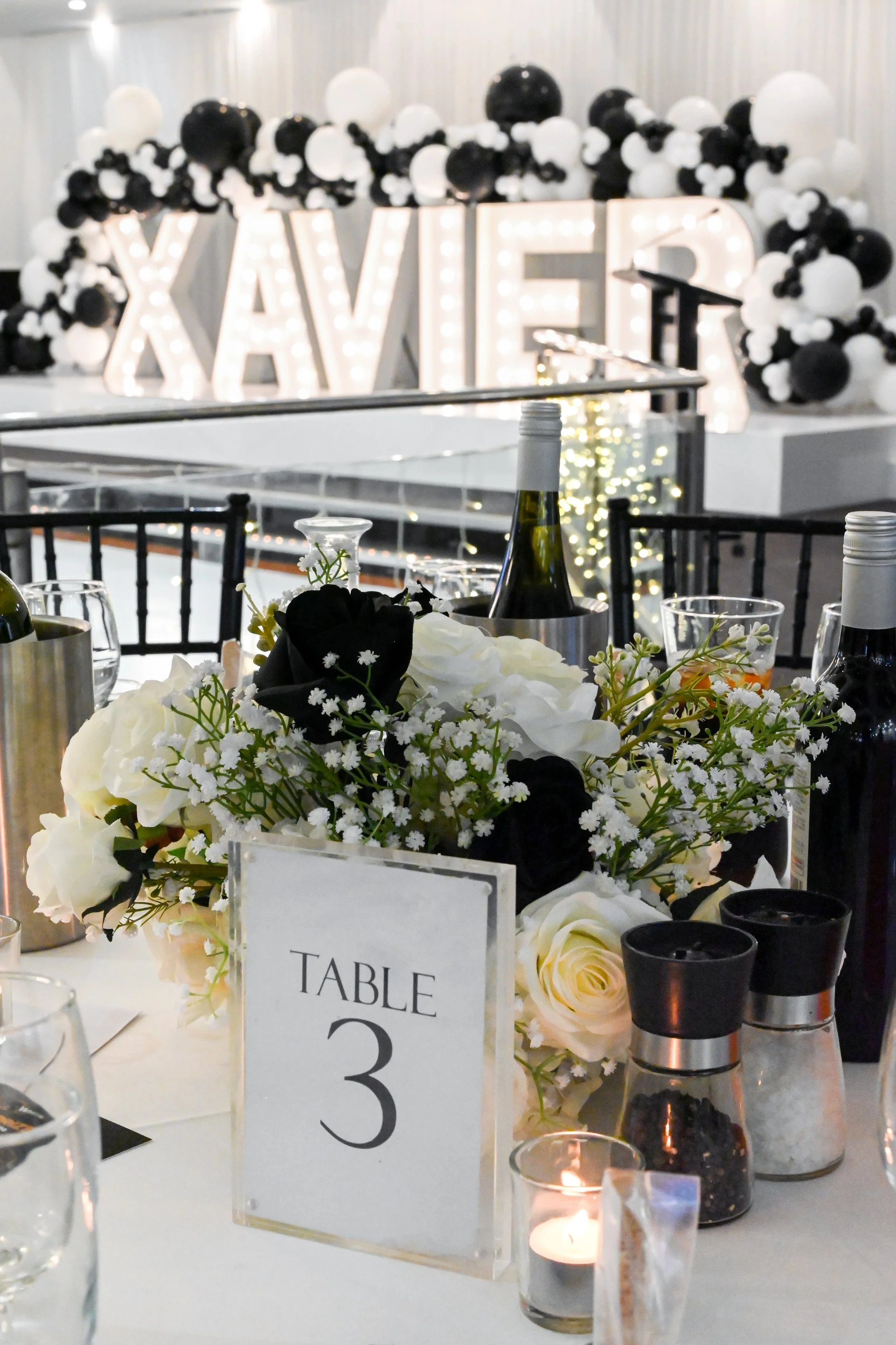Table decorated with white and black flowers, candles, and a table number sign, with a large illuminated sign reading 'XAVE' and balloon arrangements in the background.