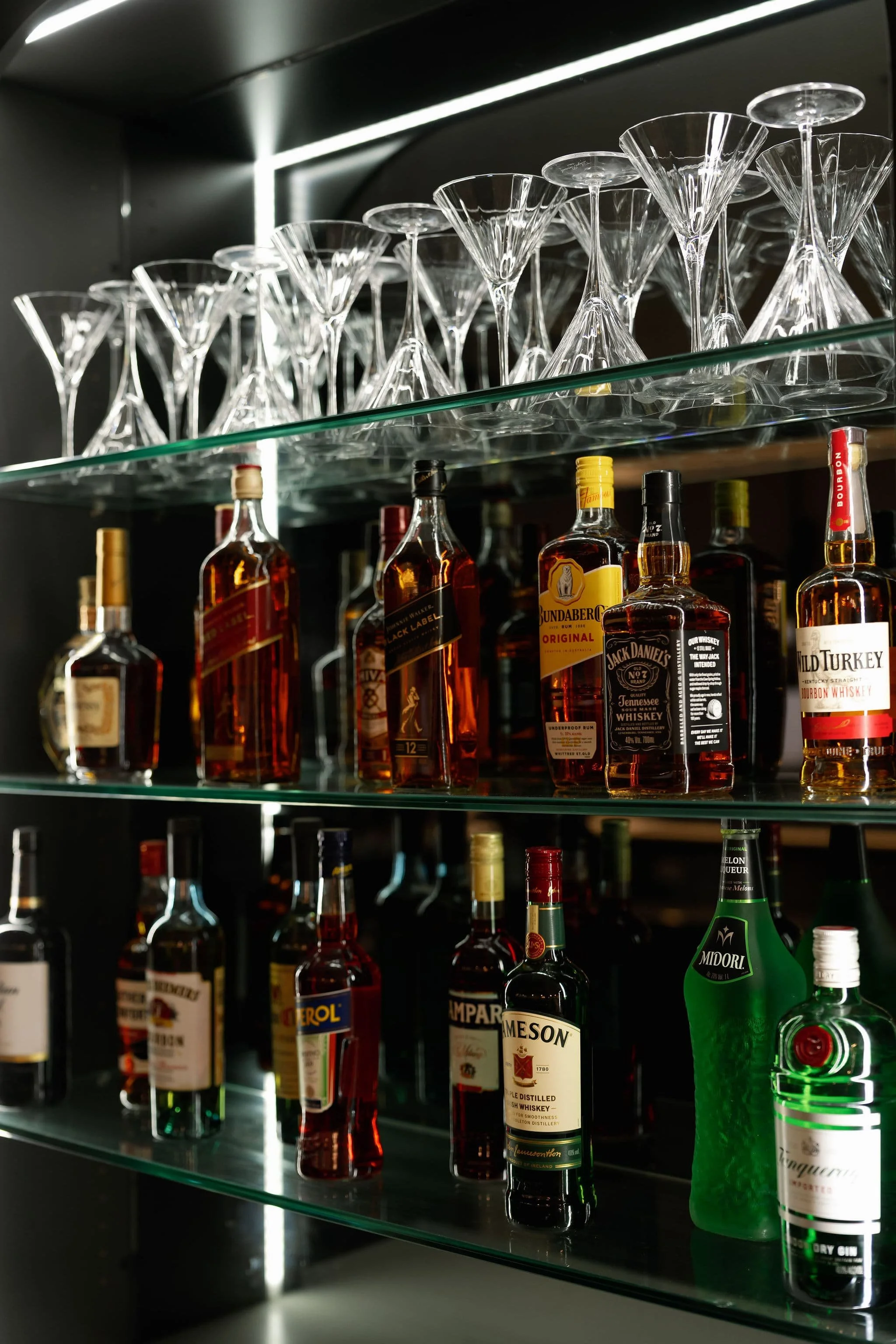 Glassware and bottles of alcohol, including whiskey, on glass shelves in a bar or liquor cabinet, with a modern dark interior design.