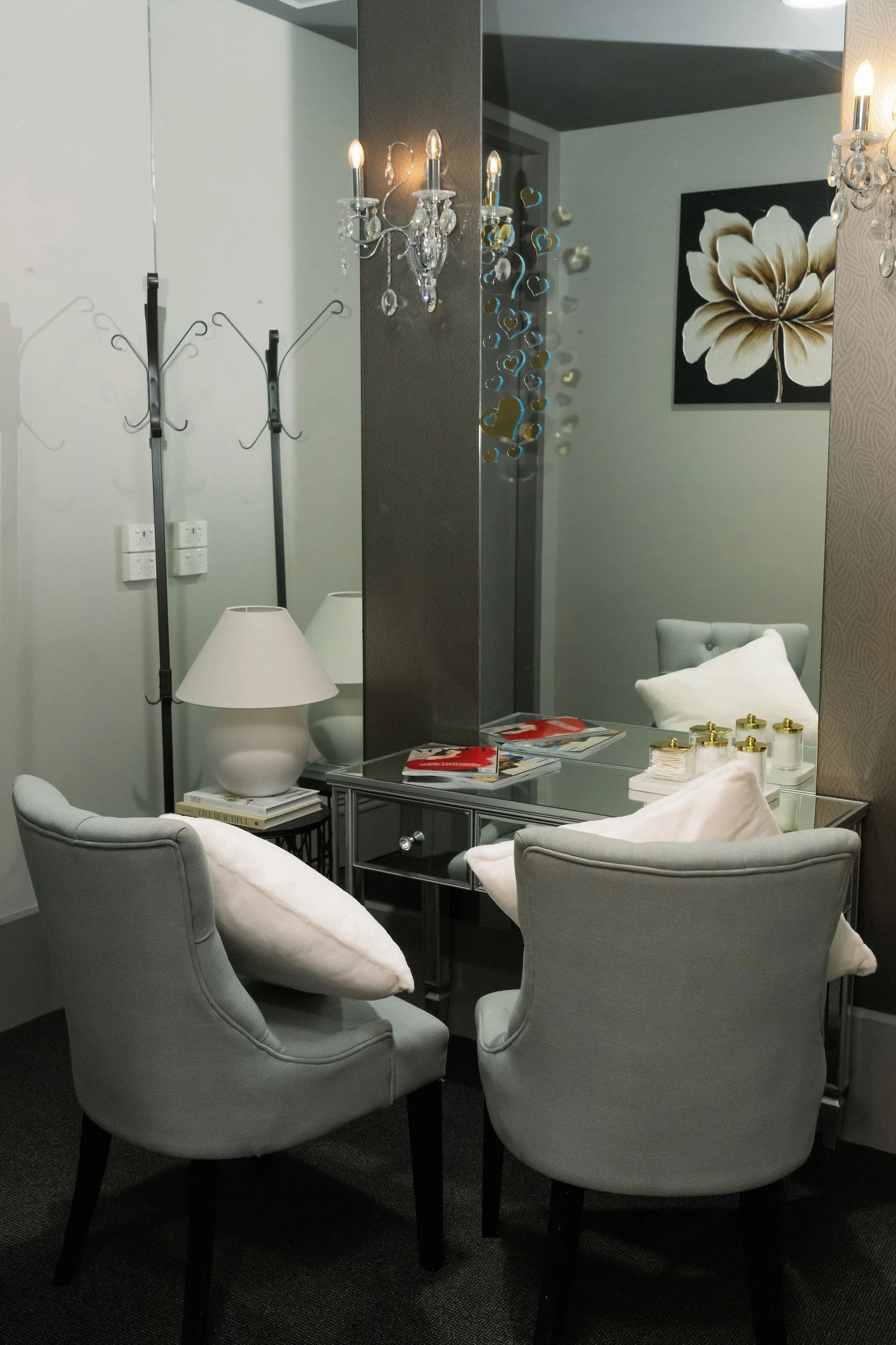 A cozy seating area with four light gray upholstered armchairs arranged around a black coffee table, topped with magazines, candles, and decorative items; a large white ceramic lamp on a side table; a mirror with a reflection of a chandelier, a wall with art of a large cream-colored flower, and a chandelier with hanging crystals.