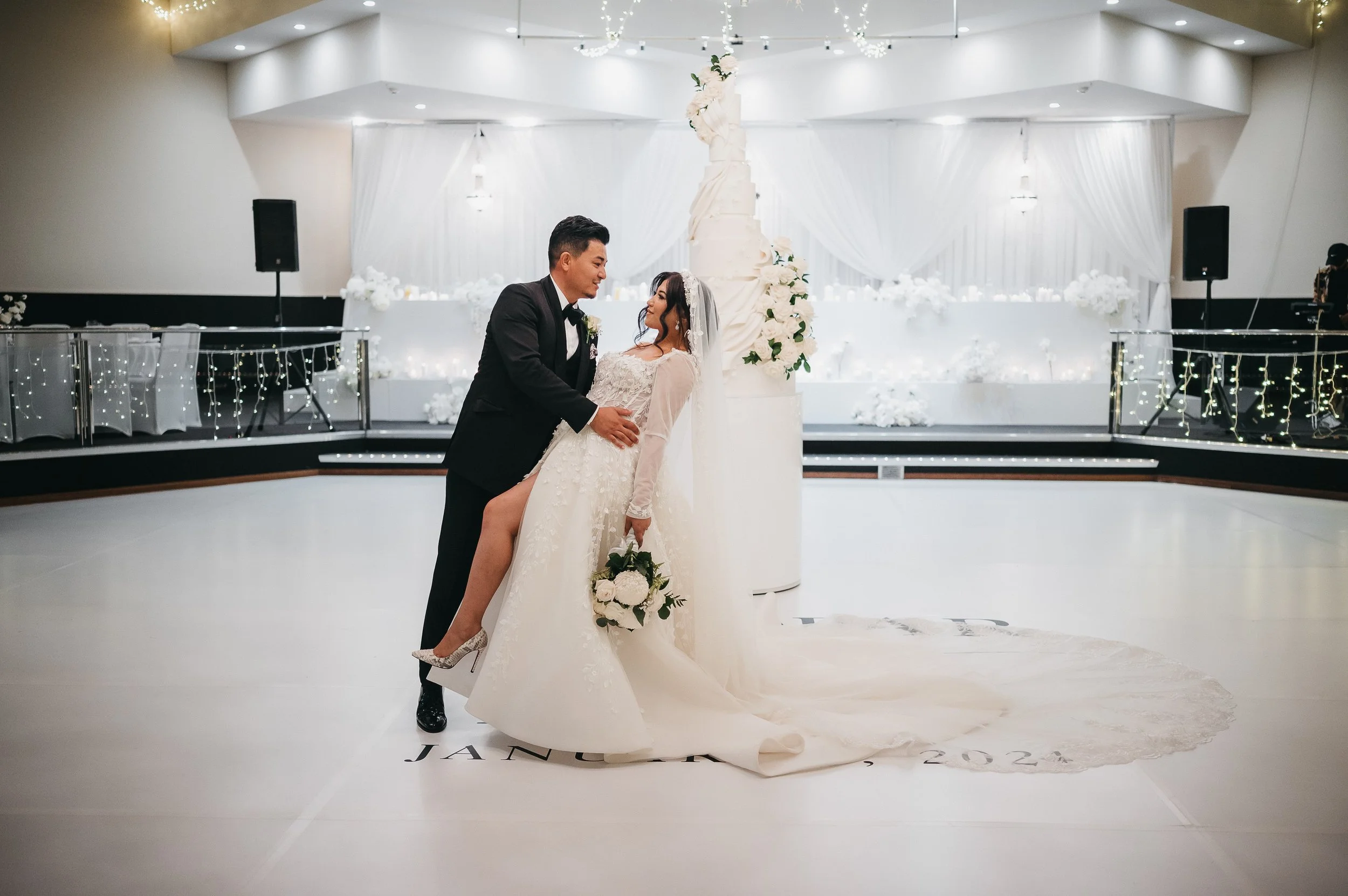 A newlywed couple dancing at their wedding reception  at Donato the venue, standing in front of a large white wedding cake adorned with flowers, in a decorated banquet hall with white drapes and string lights.