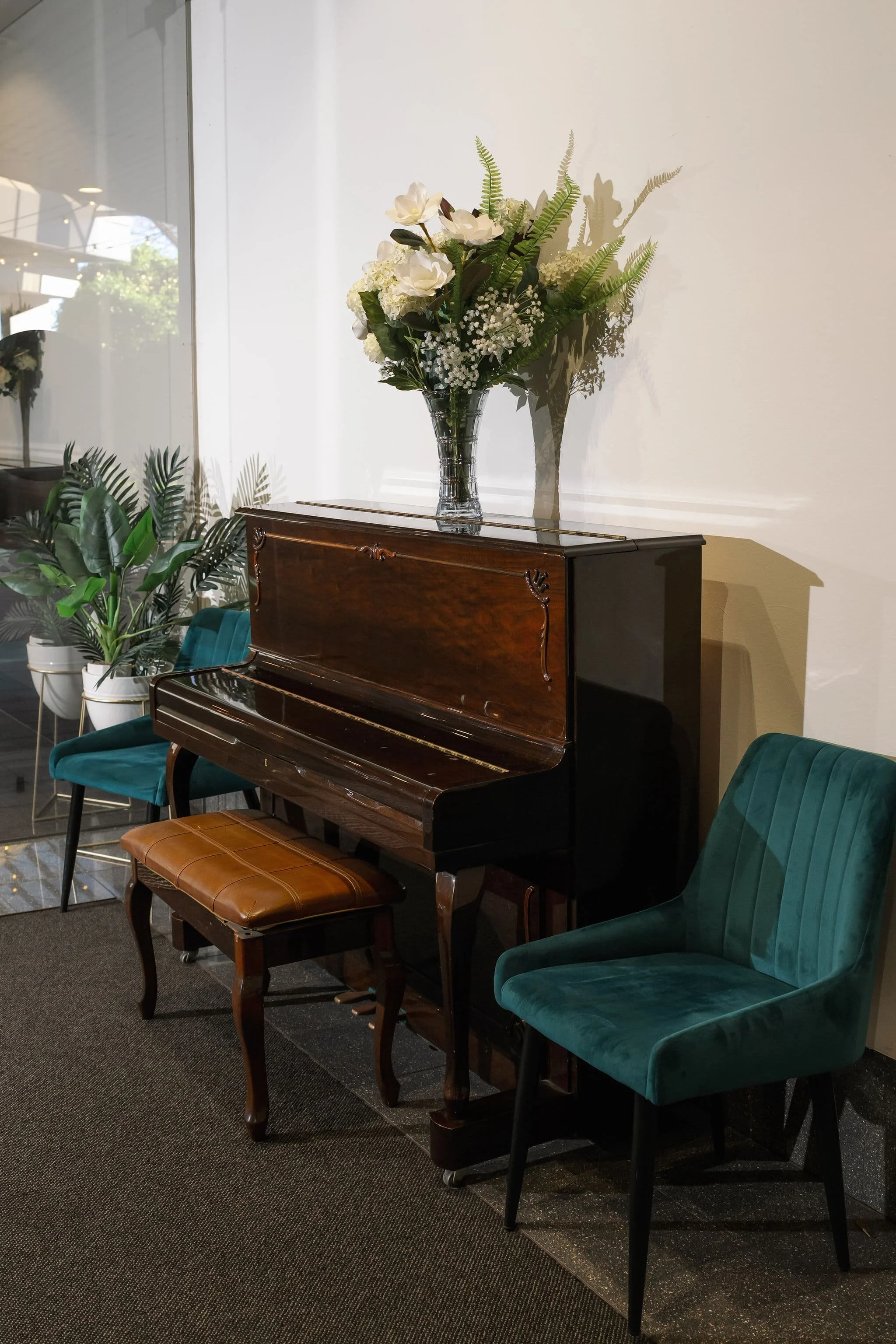 Interior of a room featuring a upright piano with wooden finish, a small brown leather bench, and a teal velvet armchair. A glass vase with white flowers and green foliage is placed on top of the piano, casting shadows on the plain white wall behind.
