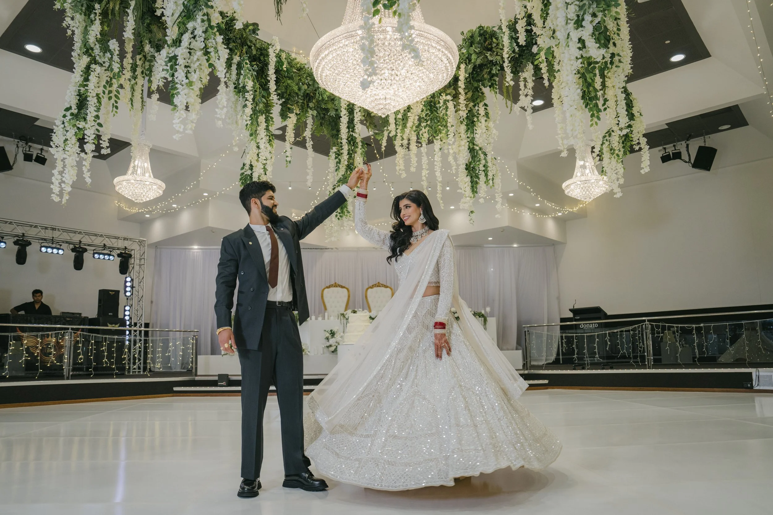 A bride and groom dancing at their wedding reception under a decorated ceiling with chandeliers and hanging floral arrangements.