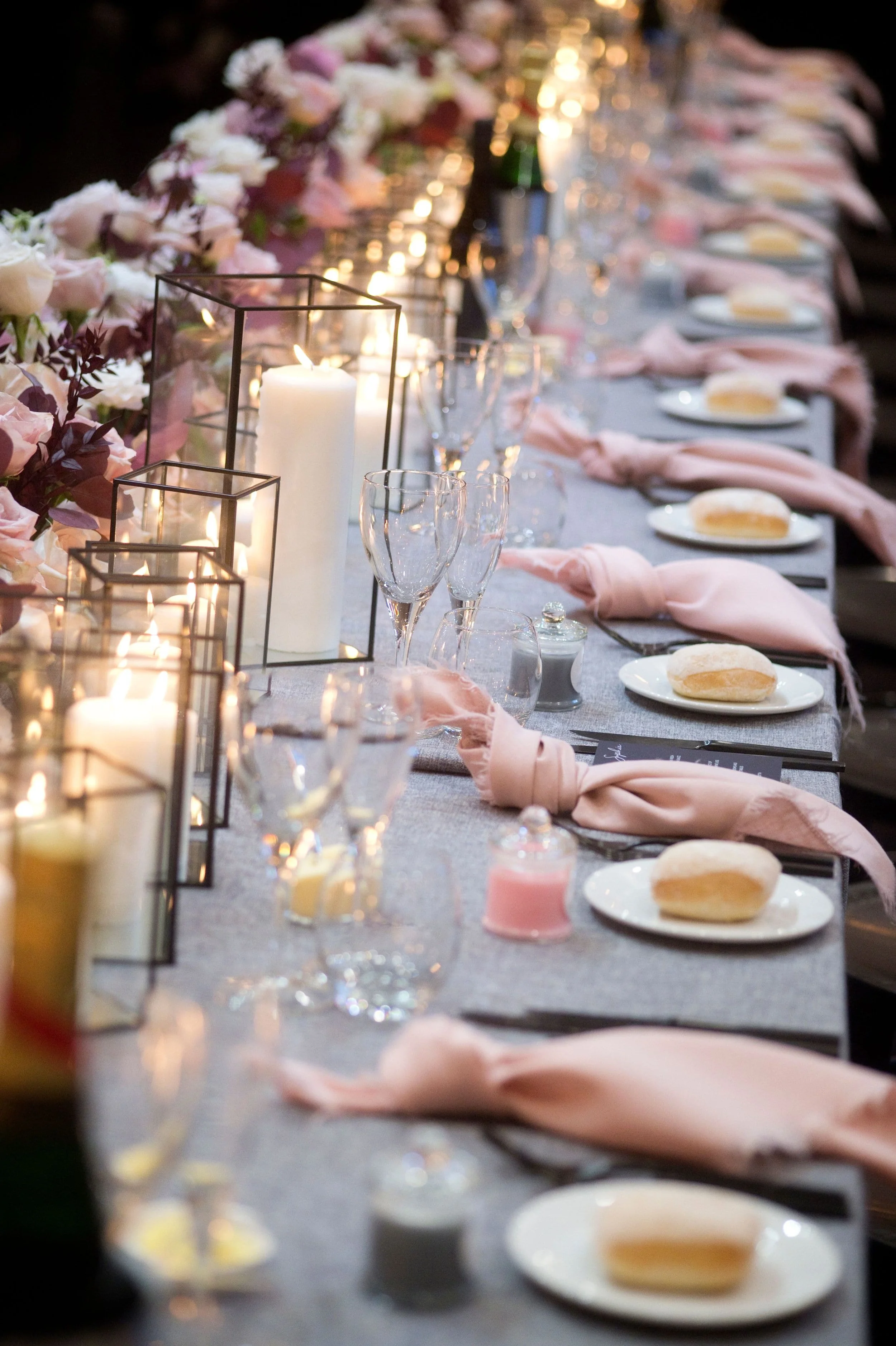 A long banquet table decorated with pink flowers, candles, glasses, and plates with bread, set for a formal event.
