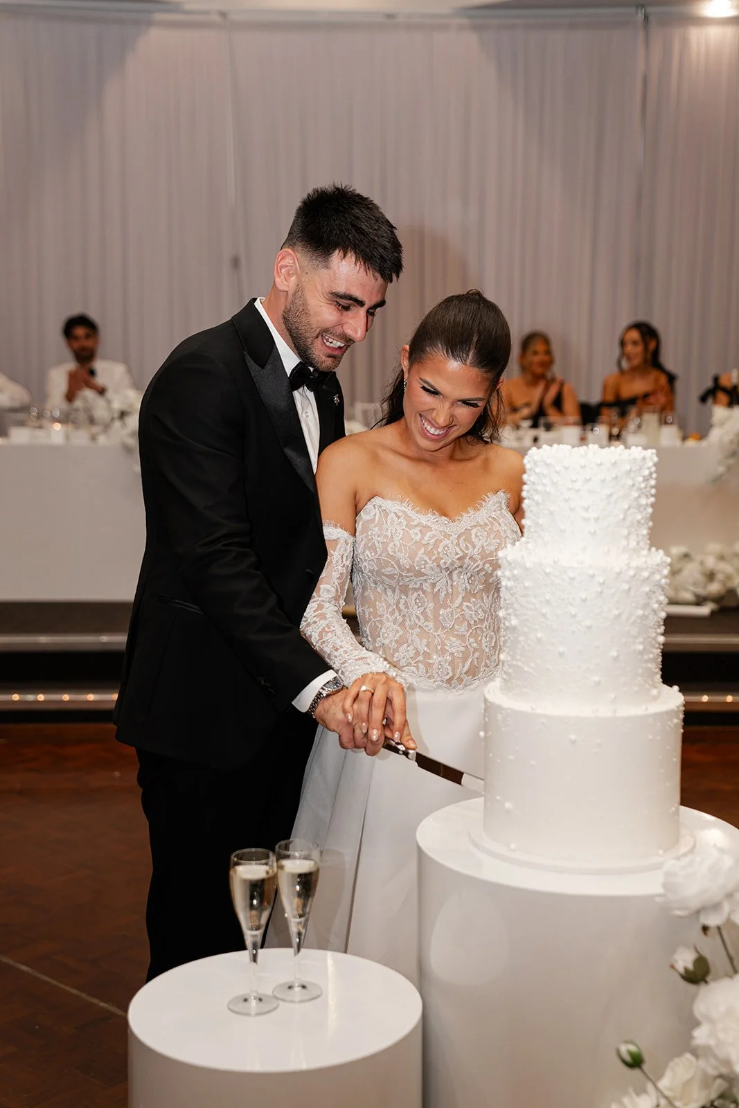 A newlywed couple in wedding attire cutting a four-tiered white wedding cake at their reception, smiling and celebrating at Donato reception centre in Adelaide