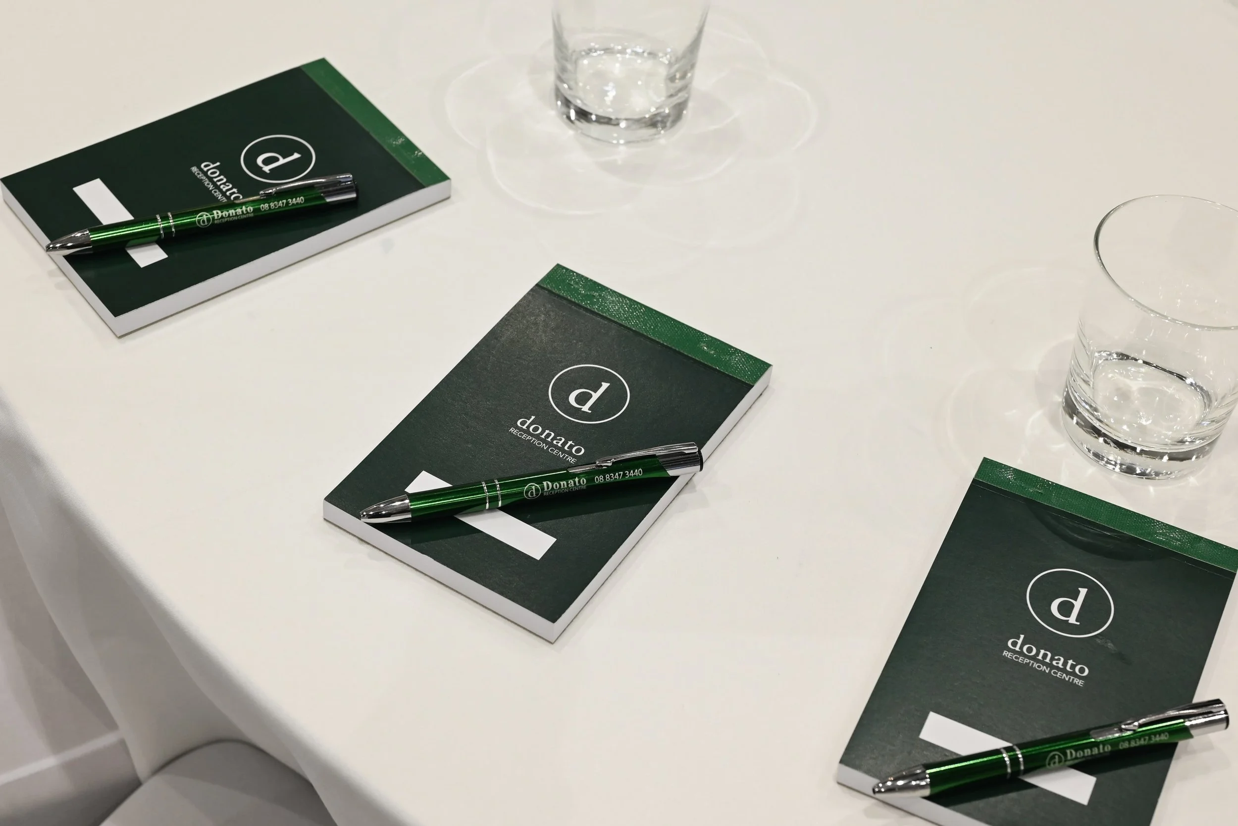 Table with green notebooks titled 'donato' and 'RECEPTION CENTRE', black pens, and glasses of water.
