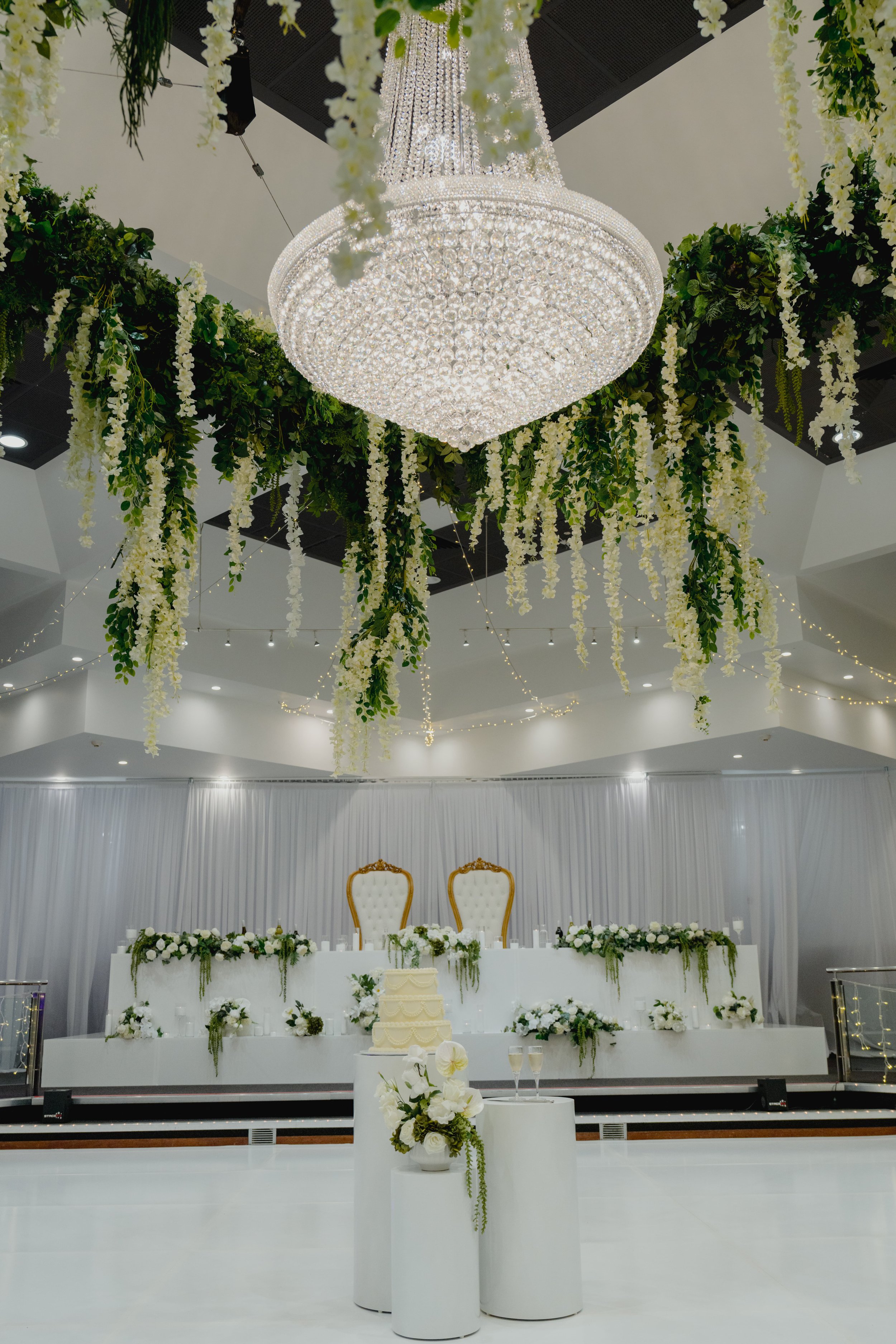 Elegant wedding reception hall with a large crystal chandelier, hanging white flowers, and a head table with floral arrangements and two gold-trimmed chairs.