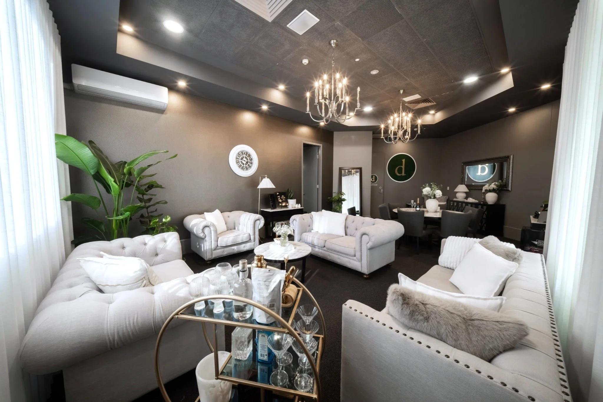 A stylish living room with white tufted sofas, a glass coffee table, potted plants, framed art, chandeliers, and a dark ceiling with recessed lighting at Donato the venue in Adelaide