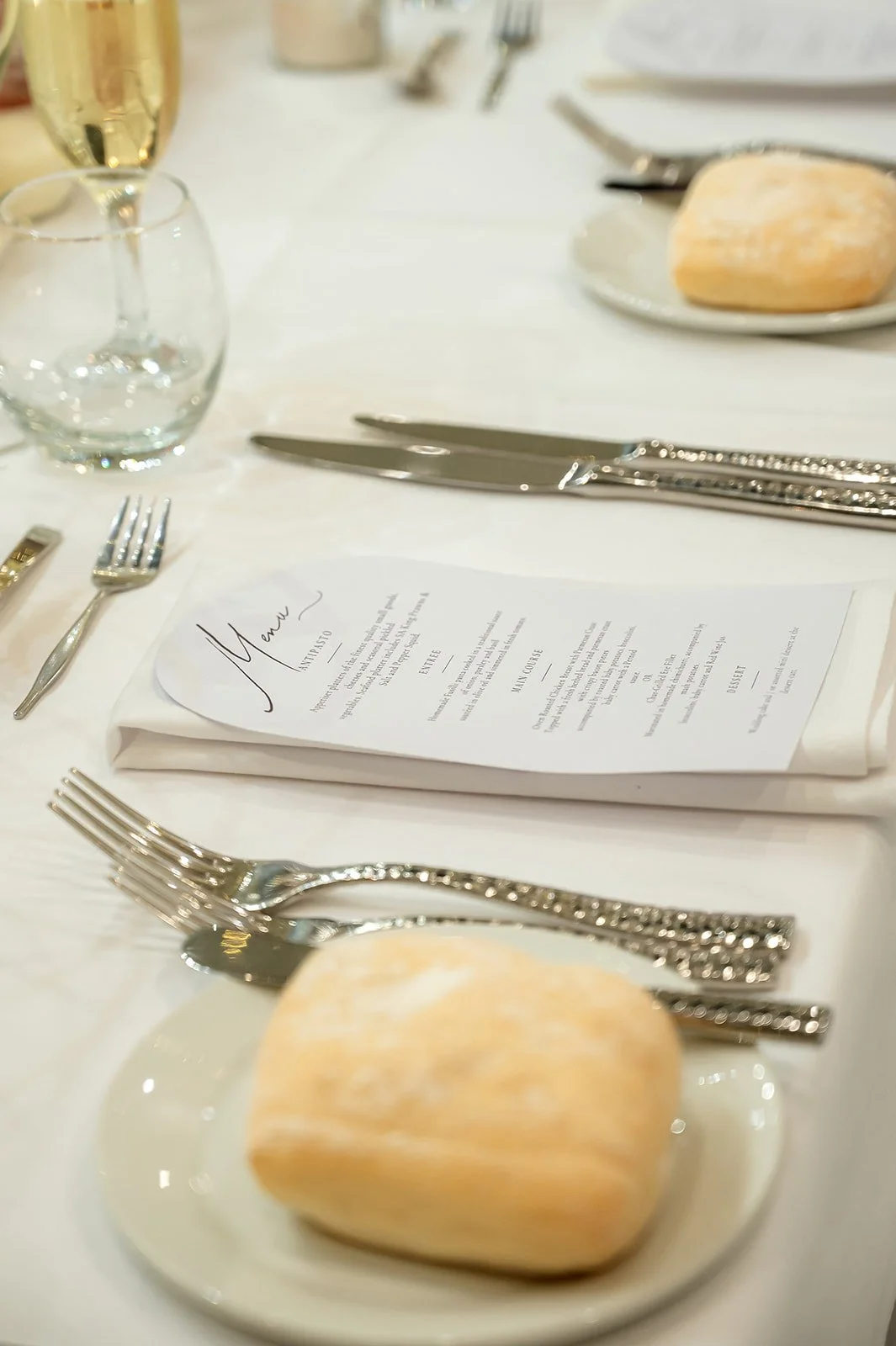 A formal table setting with a menu, silverware, a bread roll on a small plate, a glass of water, and a glass of champagne on a white tablecloth at Donato the venue in Adelaide